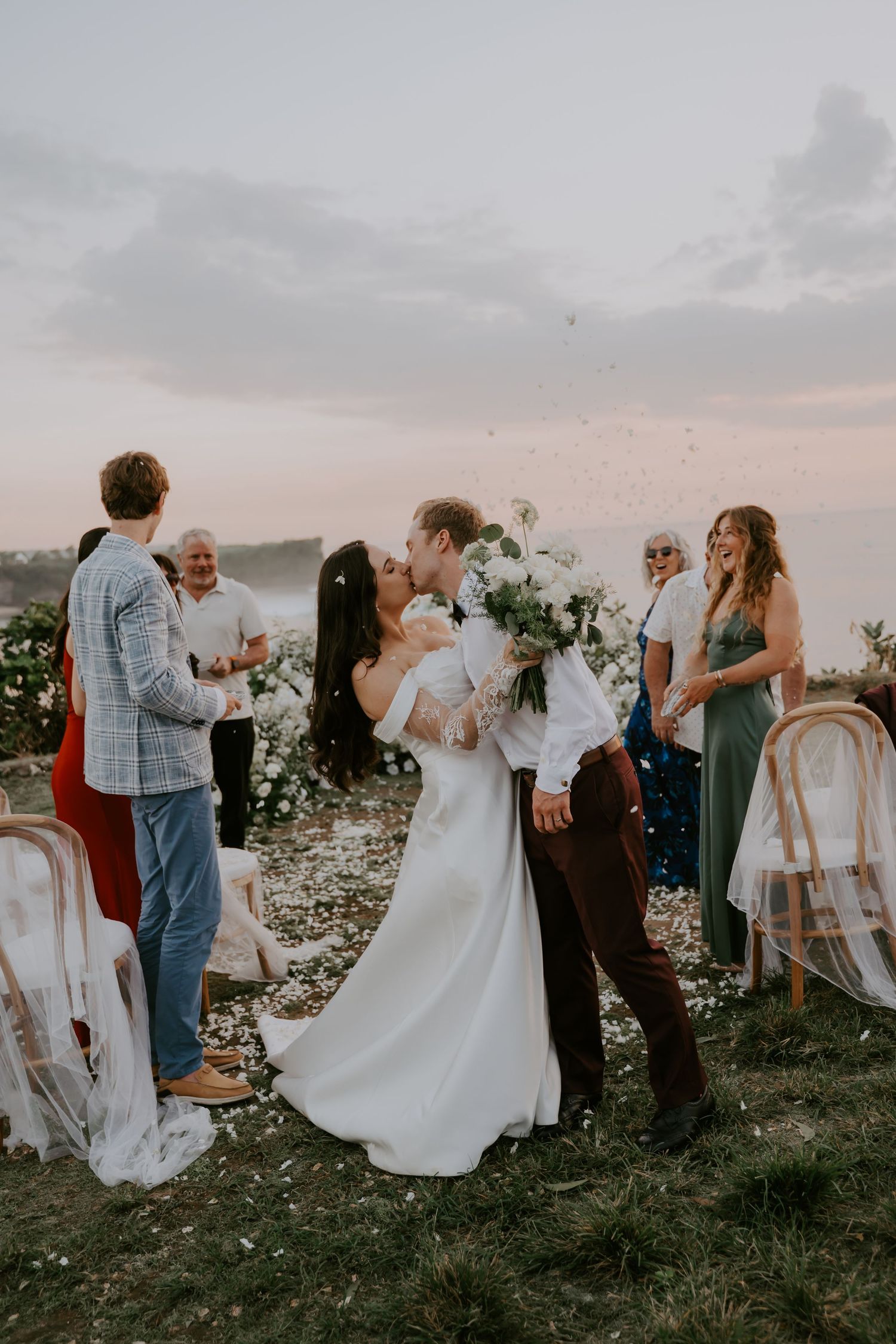 Newlyweds share first kiss as guests celebrate with confetti at sunset ceremony.