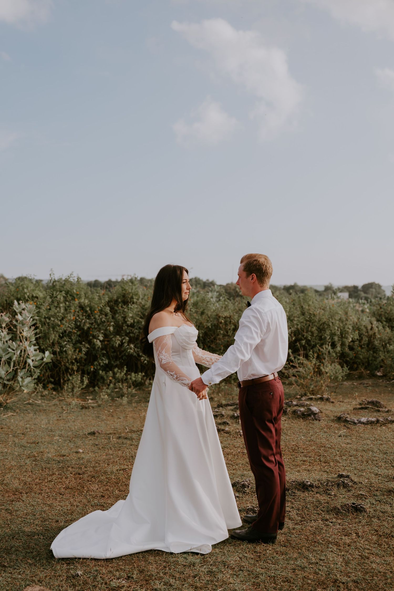 Bride in white off-shoulder gown holds hands with groom in burgundy trousers outdoors.