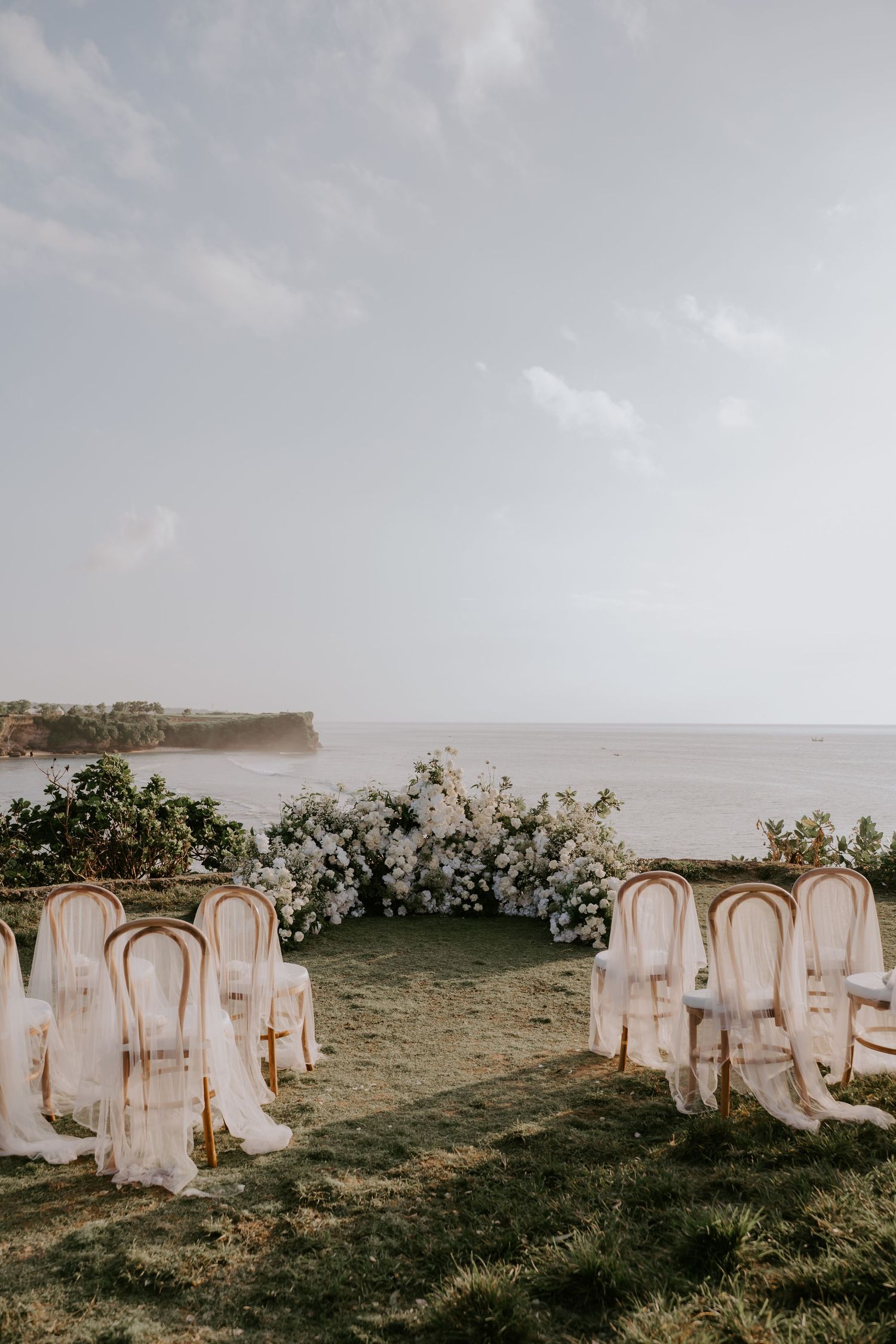 Elegant outdoor wedding ceremony setup with white floral arch and draped chairs overlooking the ocean.