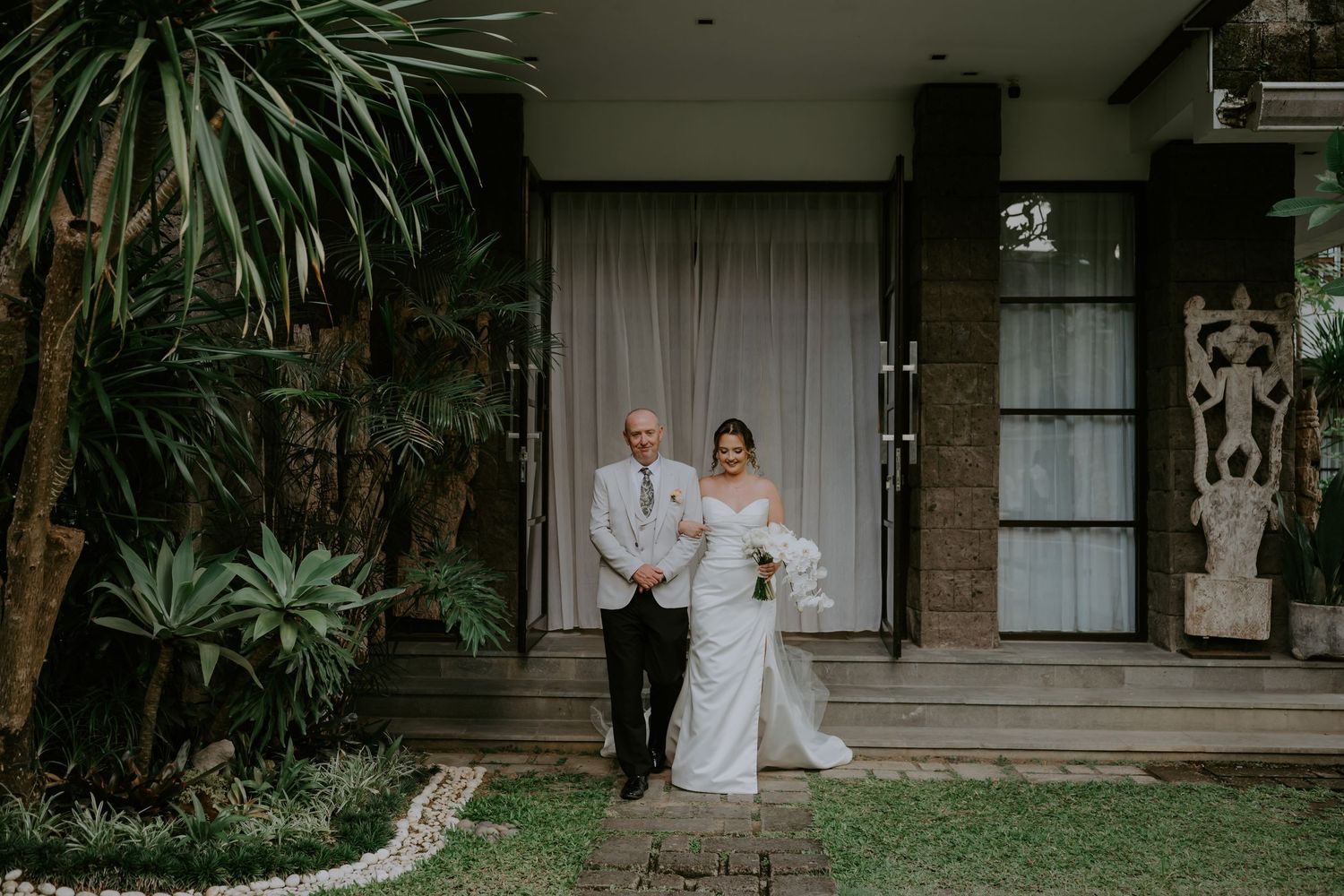 Bride walking with her father through a lush garden venue before her outdoor wedding ceremony.