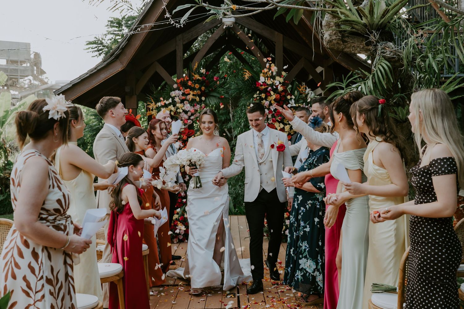Newlyweds exit ceremony as guests celebrate under a floral arch at a tropical outdoor wedding.