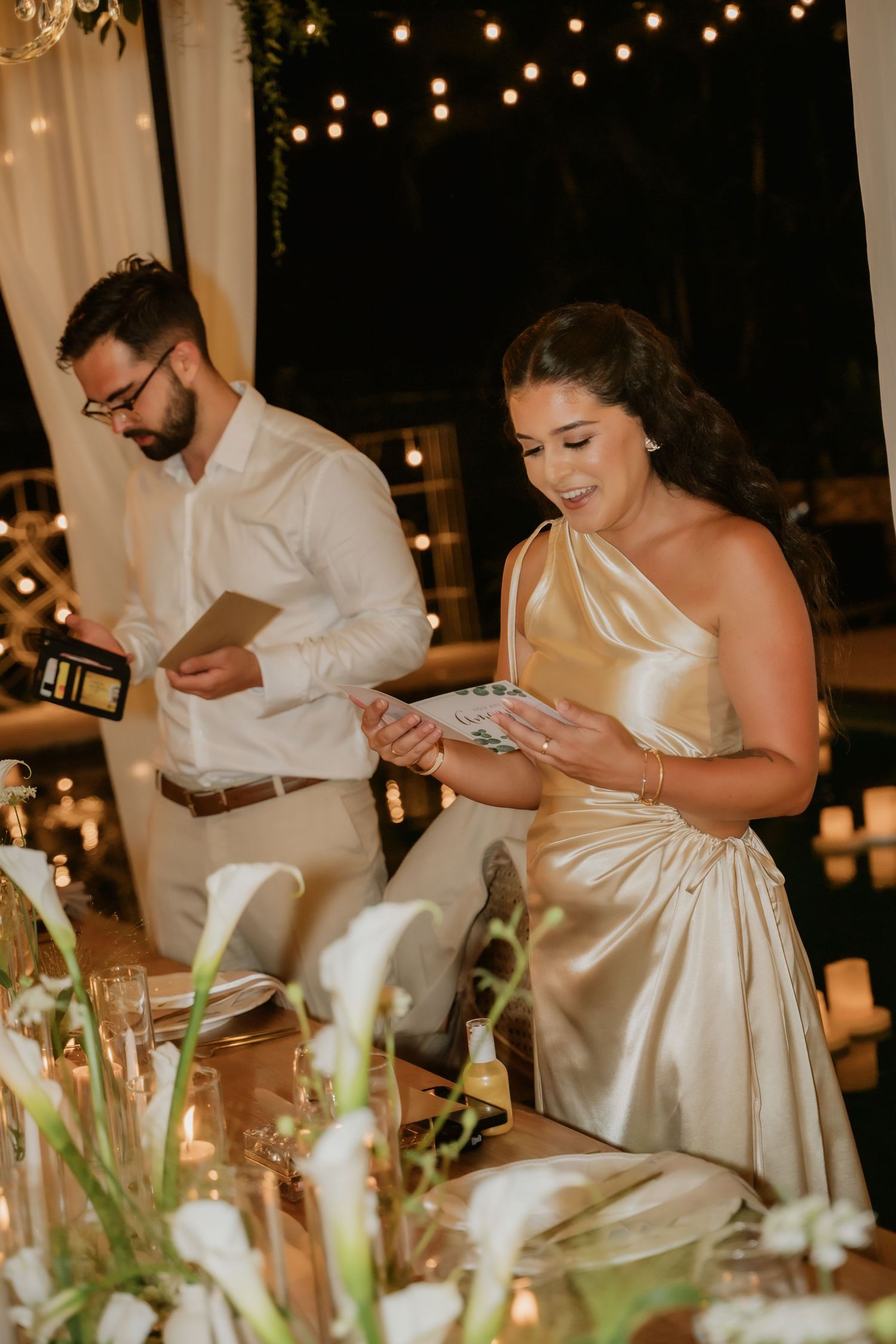 Bride in gold gown and groom in white shirt reading cards at elegant candlelit wedding reception.