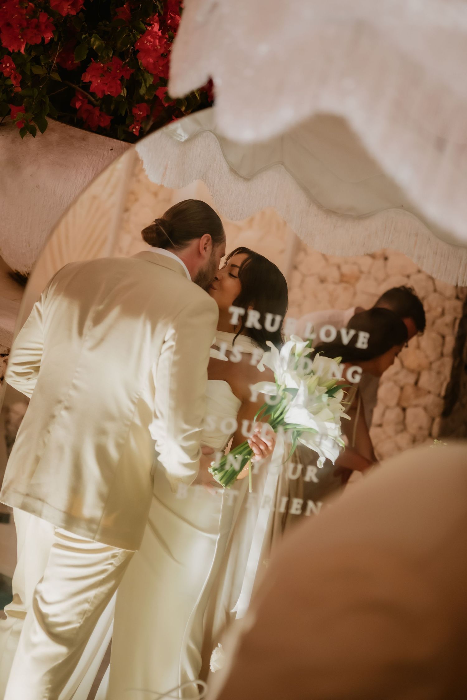 Bride and groom share romantic kiss at indoor wedding ceremony before decorative backdrop.