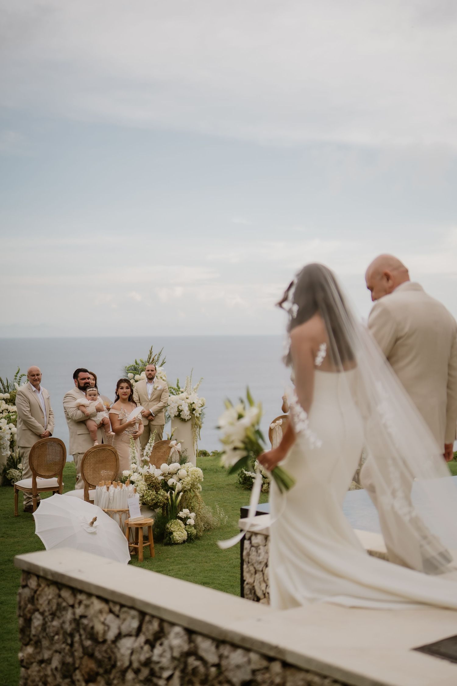 Bride in a white gown and veil walking down the aisle with her father at an oceanfront wedding ceremony.