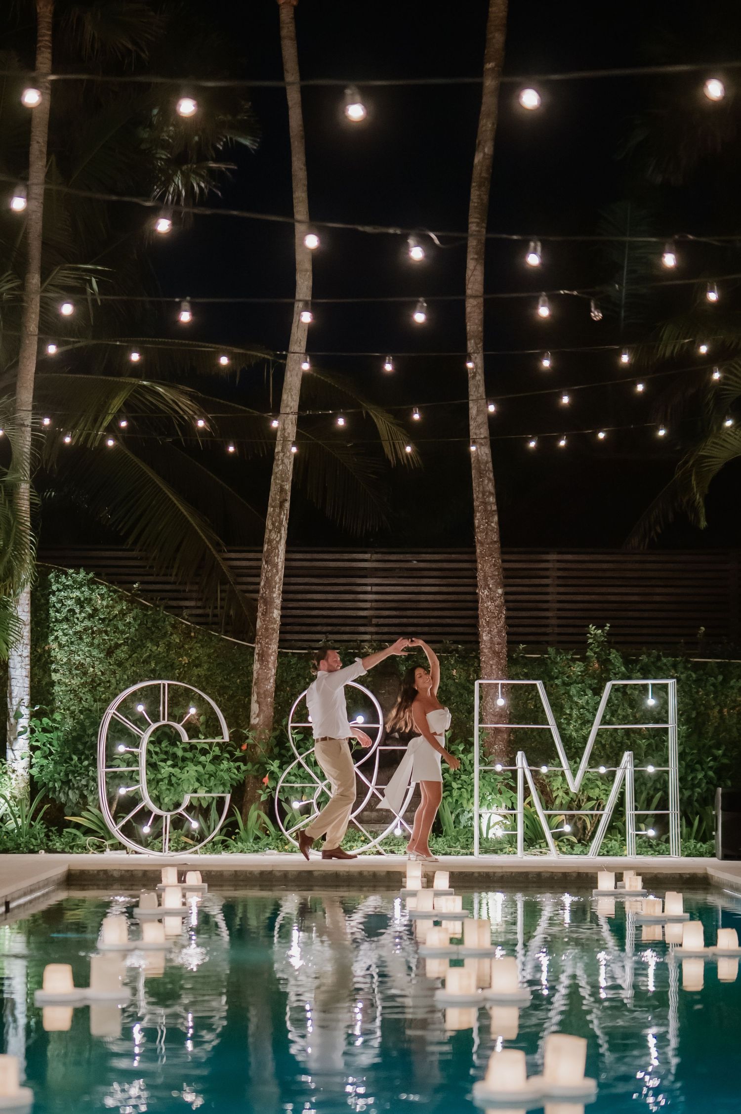 Couple dancing by pool at night with C&M marquee letters and string lights overhead.
