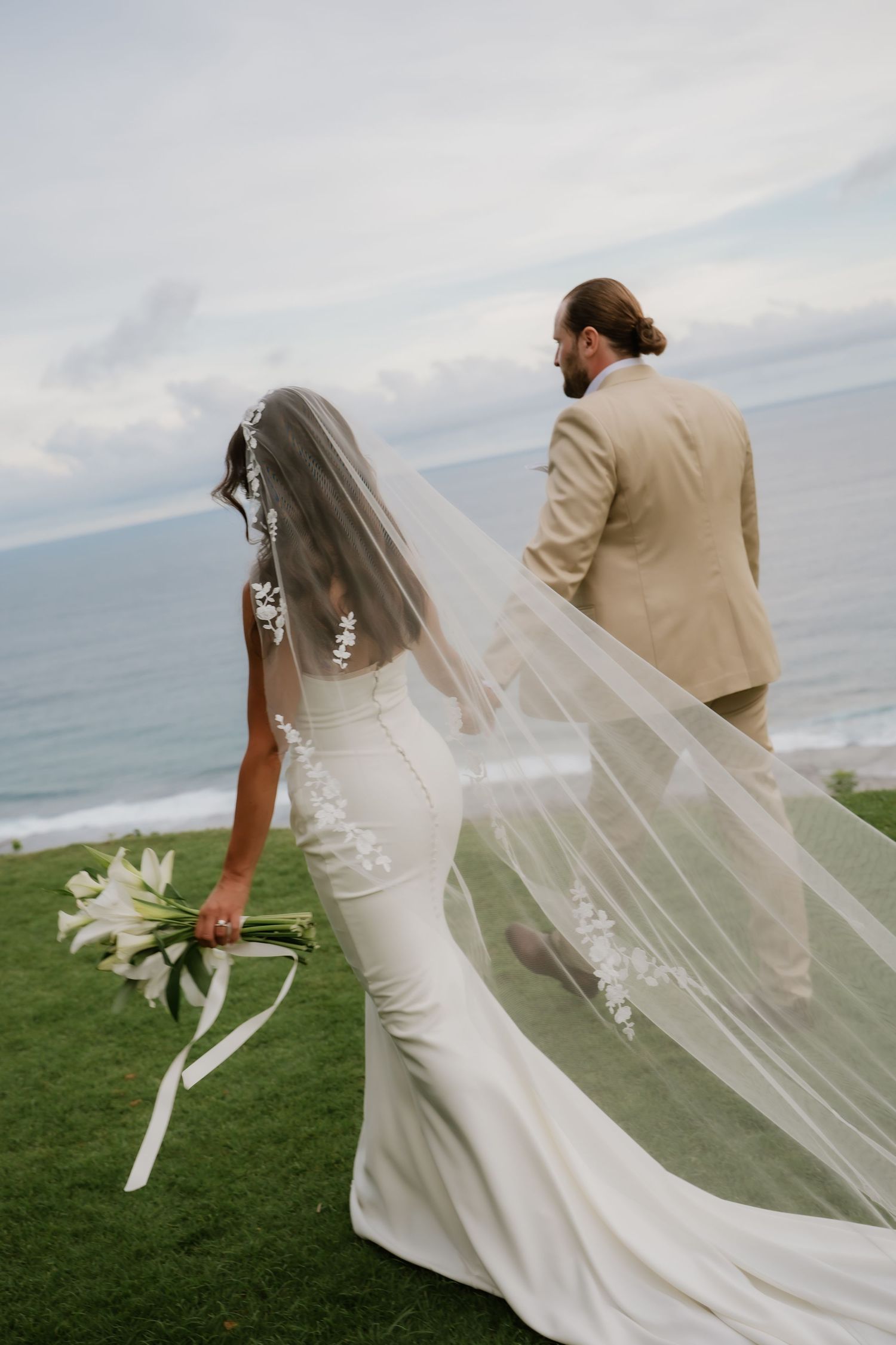 Bride and groom walking on green lawn with ocean backdrop, veil flowing in the wind at seaside wedding.