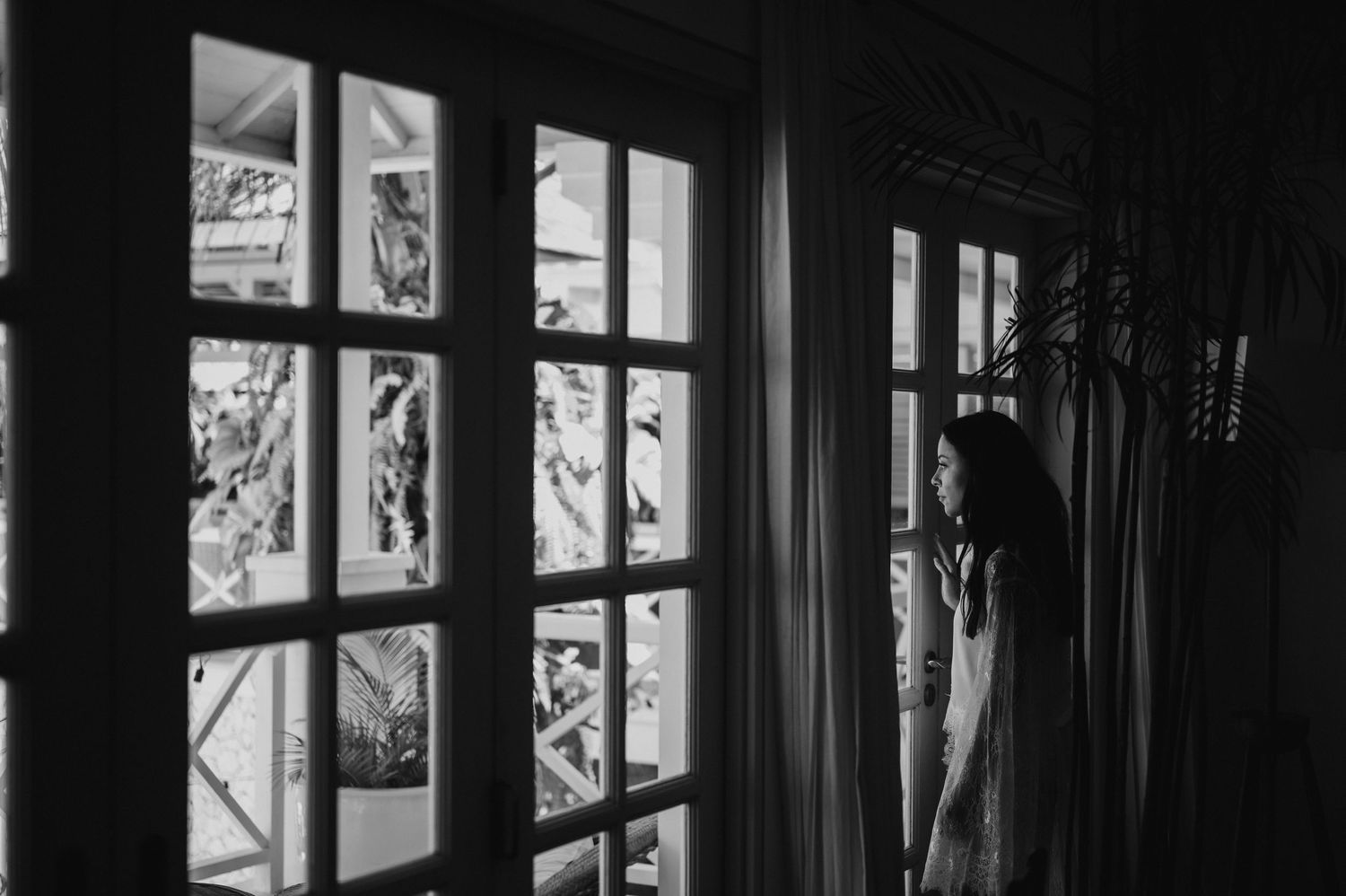 Black and white photo of a woman standing by French doors, gazing outside through sheer curtains in dim light.