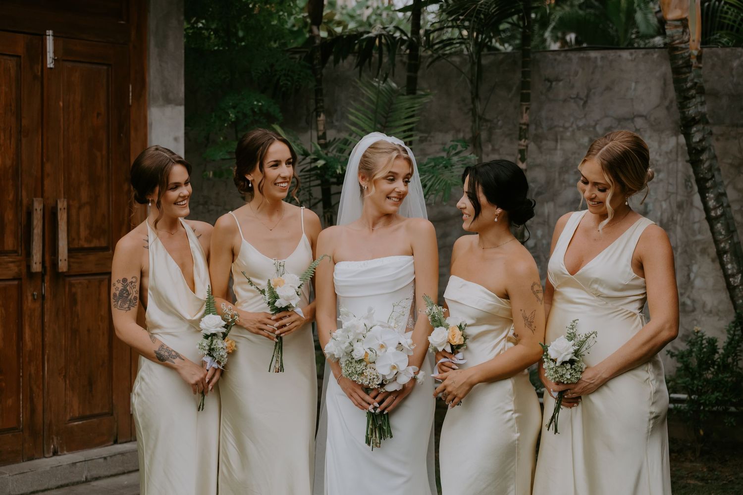 Bride in white strapless gown smiling with four bridesmaids in ivory dresses holding bouquets.