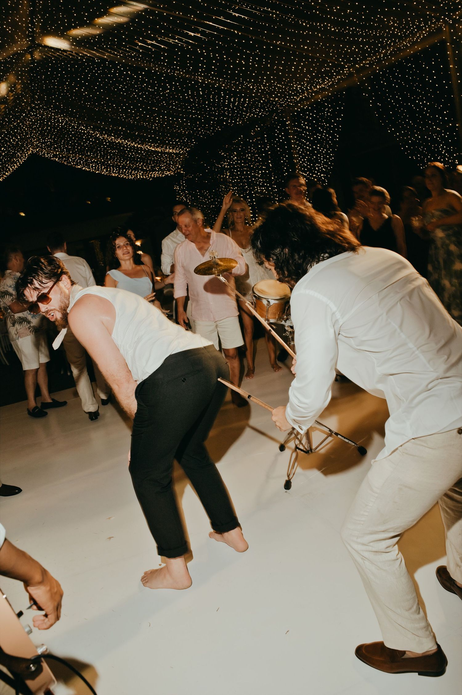 Energetic wedding guests dancing barefoot on a white dance floor under a canopy of fairy lights.