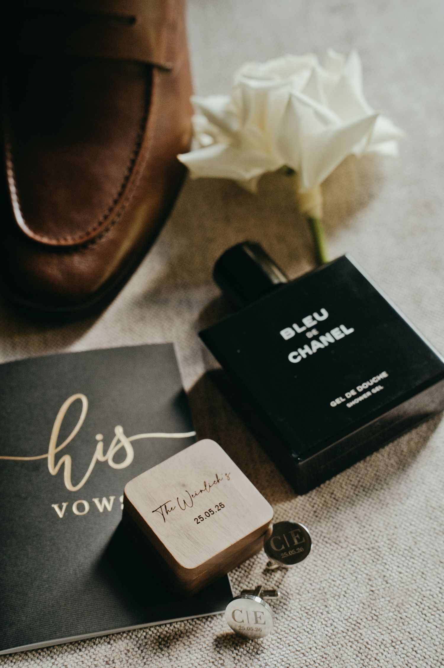 Groom wedding flat lay with Bleu de Chanel cologne, vow book, cufflinks, wooden ring box, and brown suede loafer.