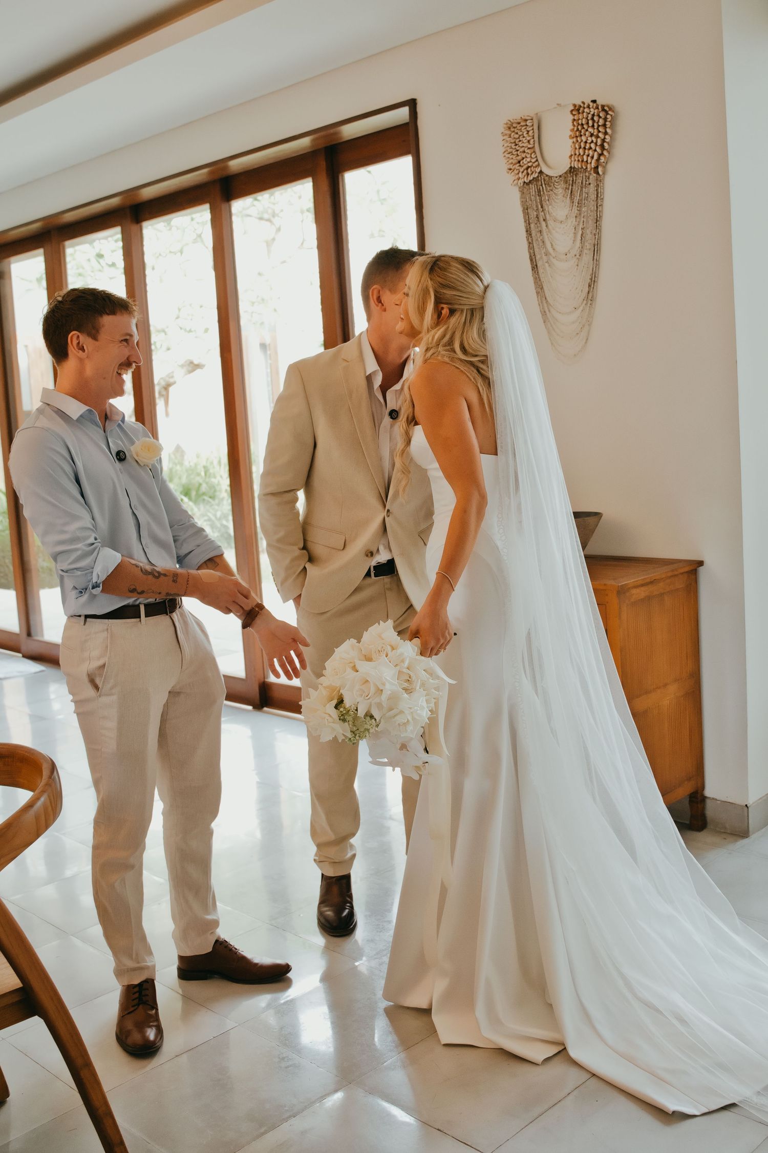 Bride and groom share a kiss while a smiling groomsman holds their hands during an intimate indoor wedding ceremony.