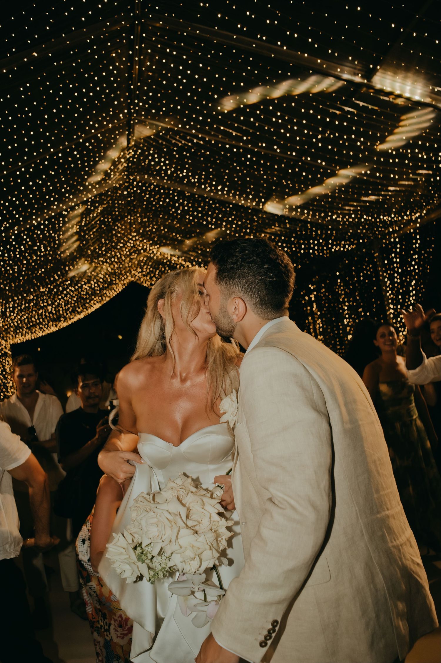 Bride and groom sharing a kiss under sparkling fairy lights at night wedding reception.