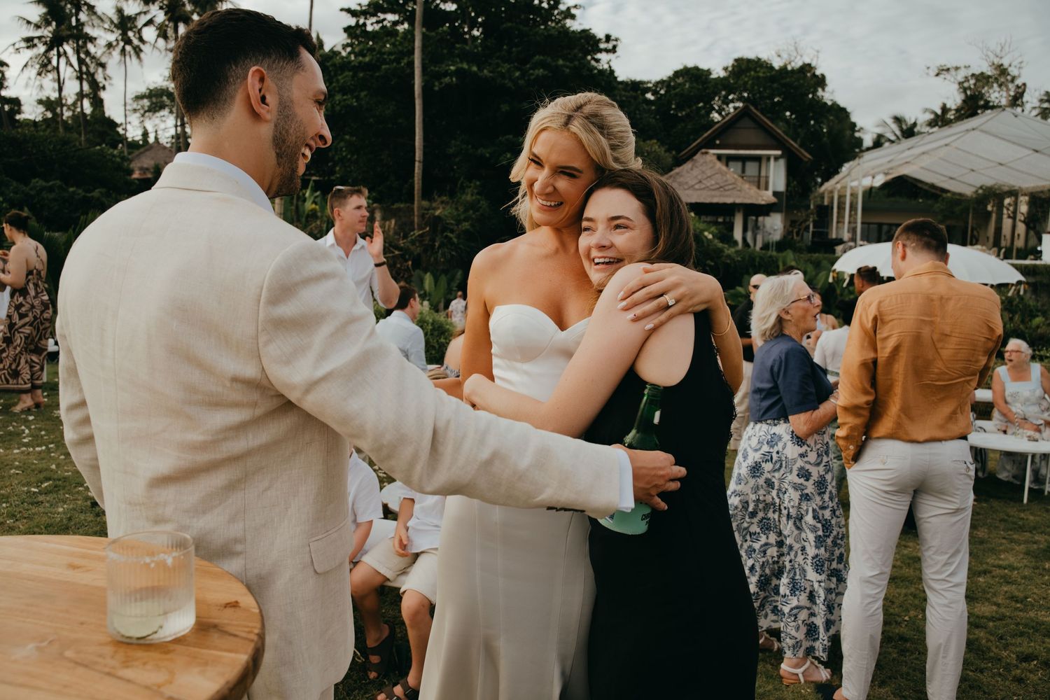 Bride in strapless gown and groom embrace a joyful guest during outdoor wedding reception.
