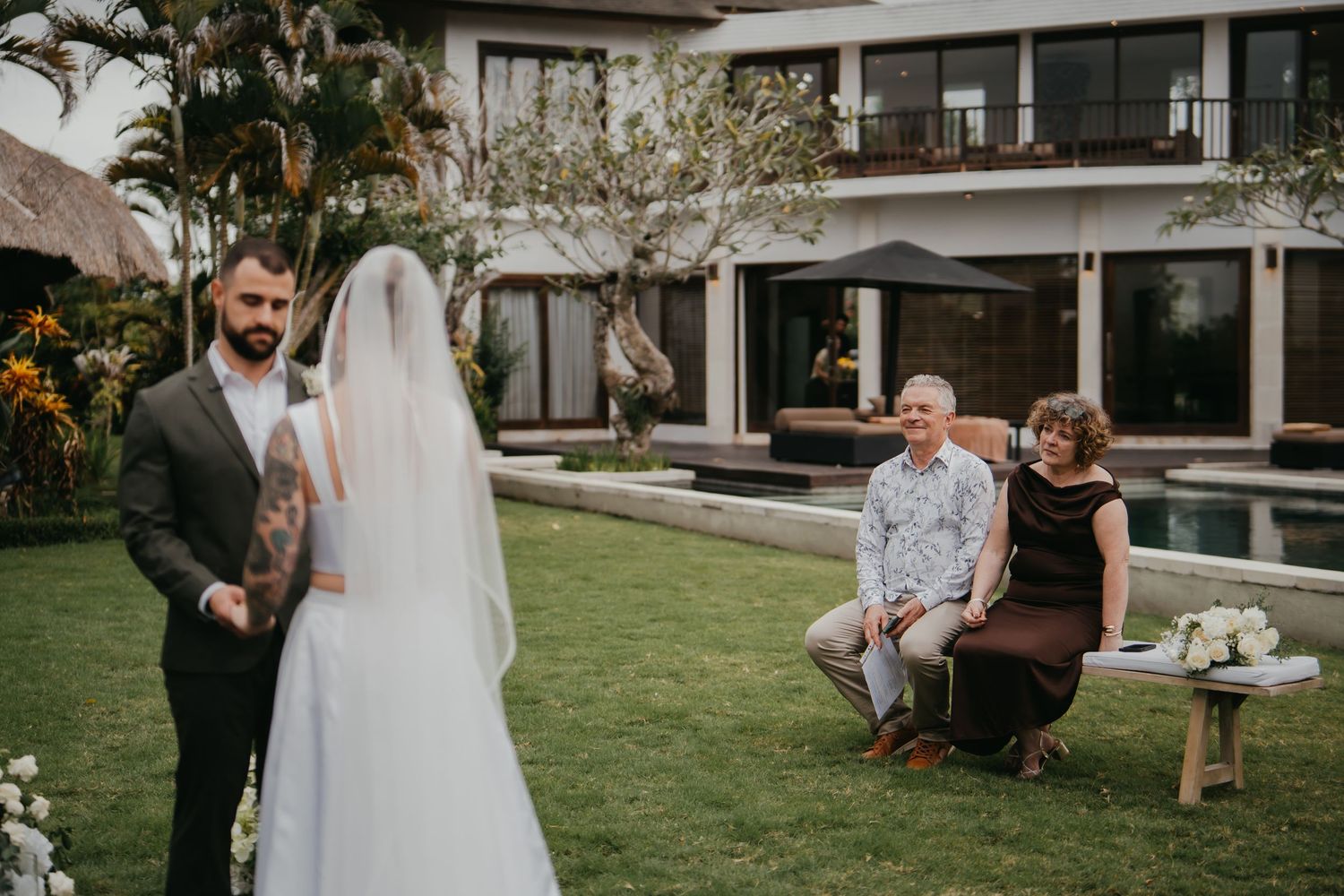 Groom lifts brides veil as seated guests smile during intimate outdoor Bali garden wedding.