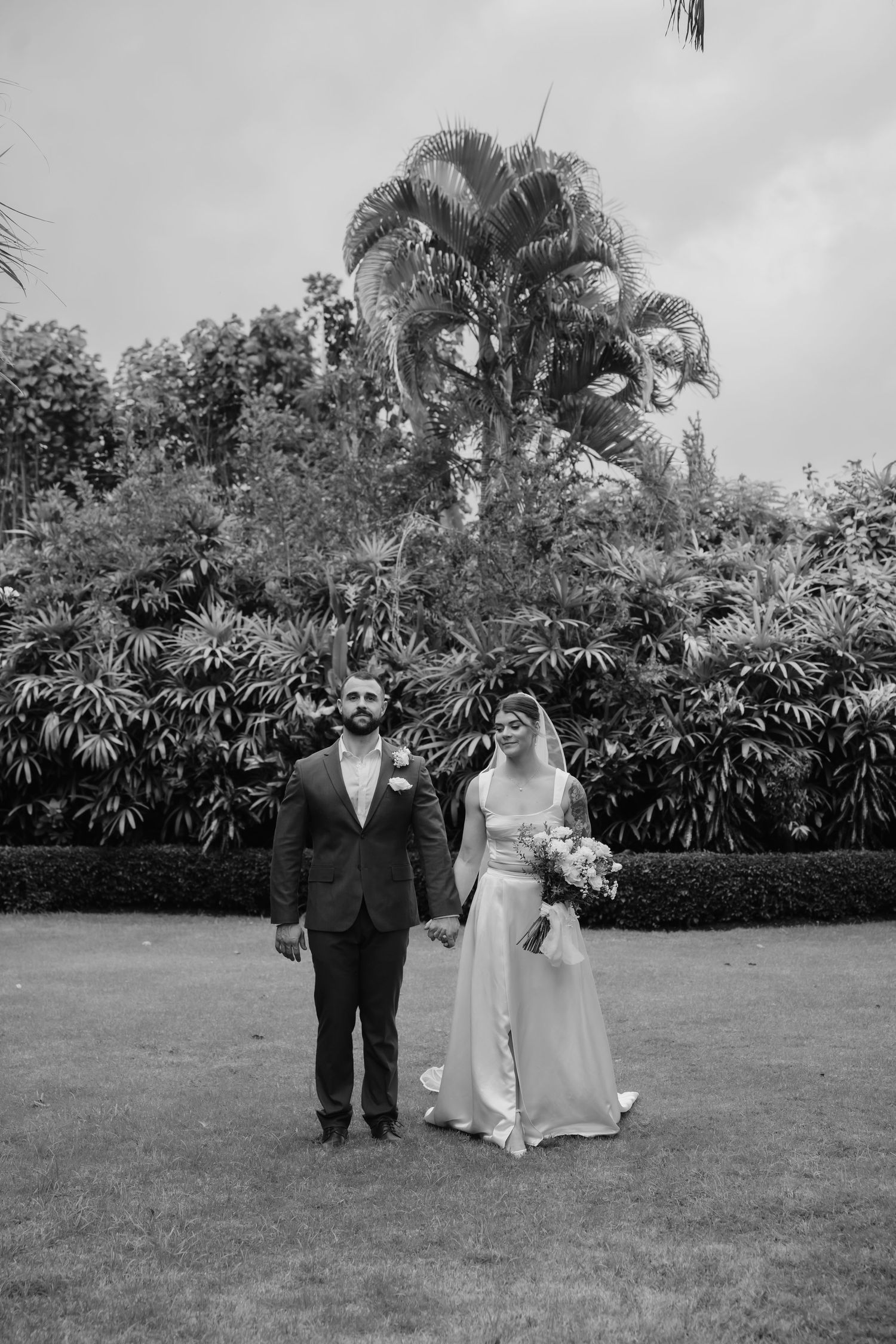 Black and white portrait of bride and groom holding hands in lush tropical garden setting.