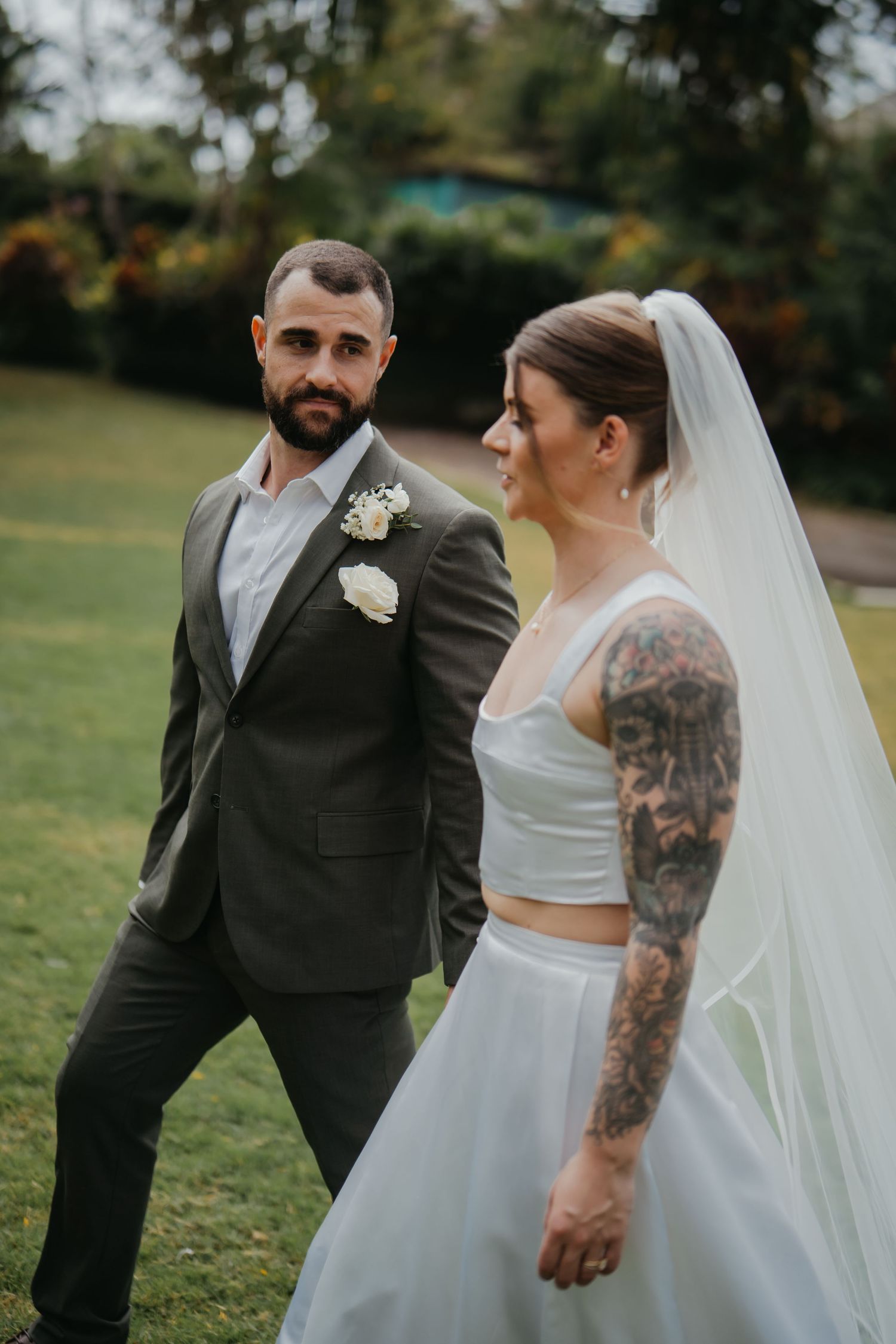 Groom in grey suit walking with tattooed bride in two-piece gown on green lawn.