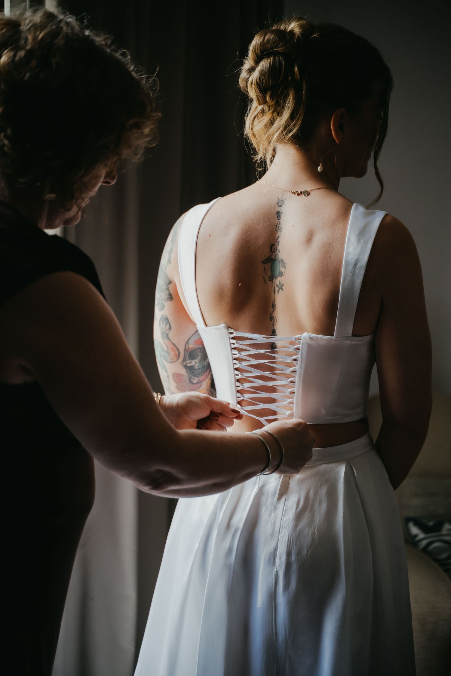 Assistant lacing up brides white corset wedding gown revealing back tattoos in dim light.