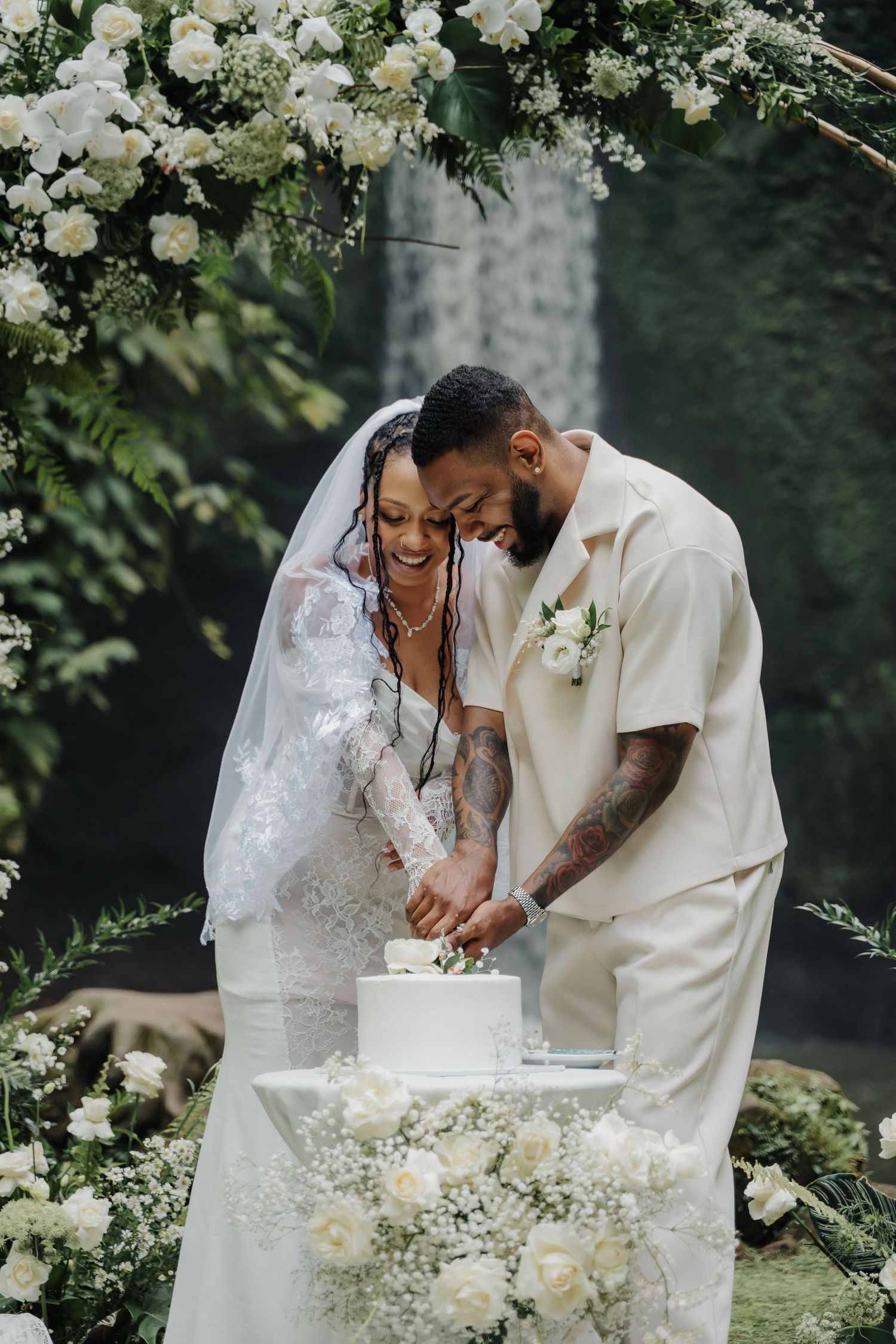 Smiling bride and groom cut a white wedding cake beneath a floral arch near a waterfall.