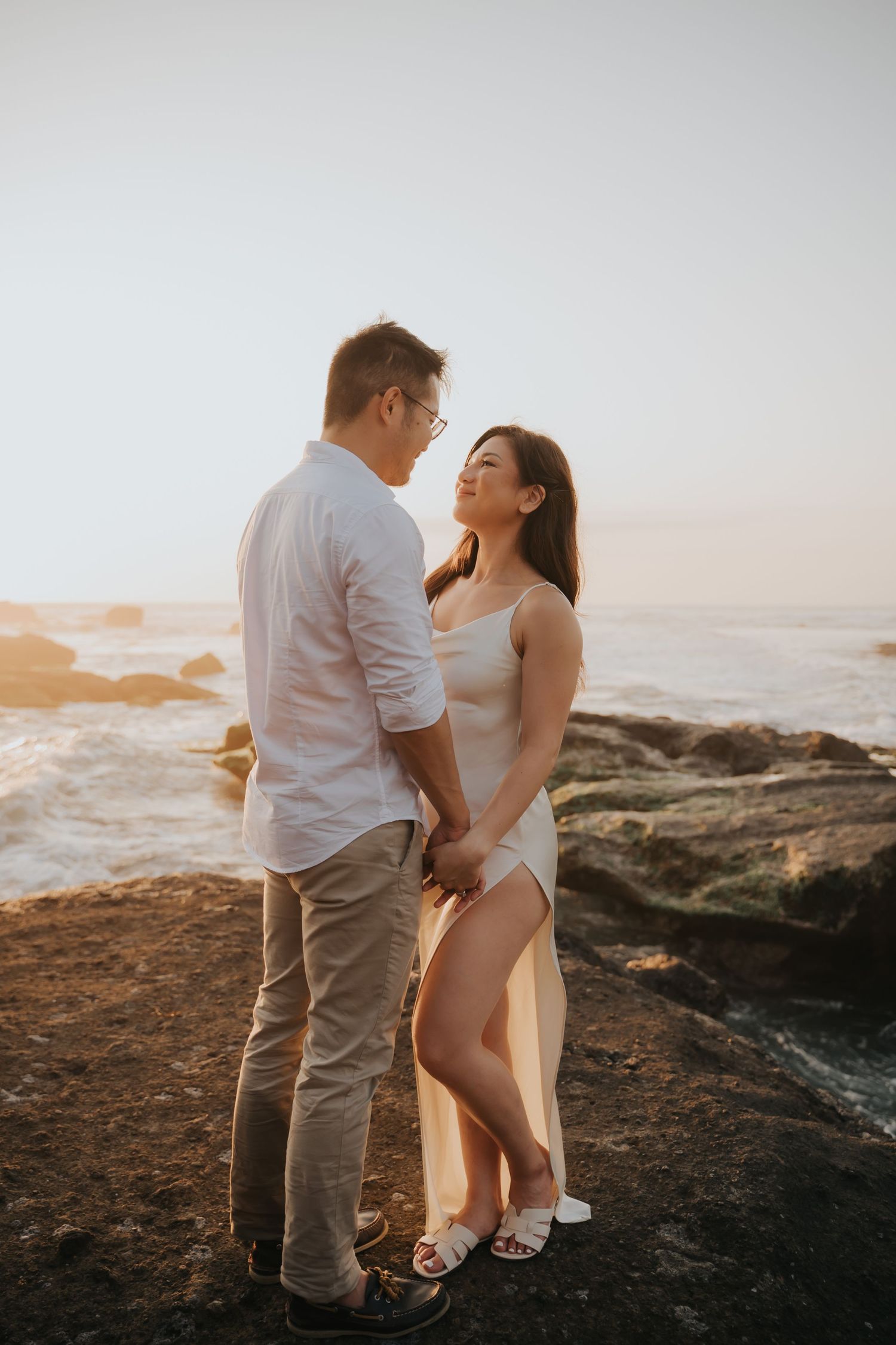 Couple holding hands on rocky coastal cliffs at golden hour sunset, sharing a romantic moment by the ocean waves.