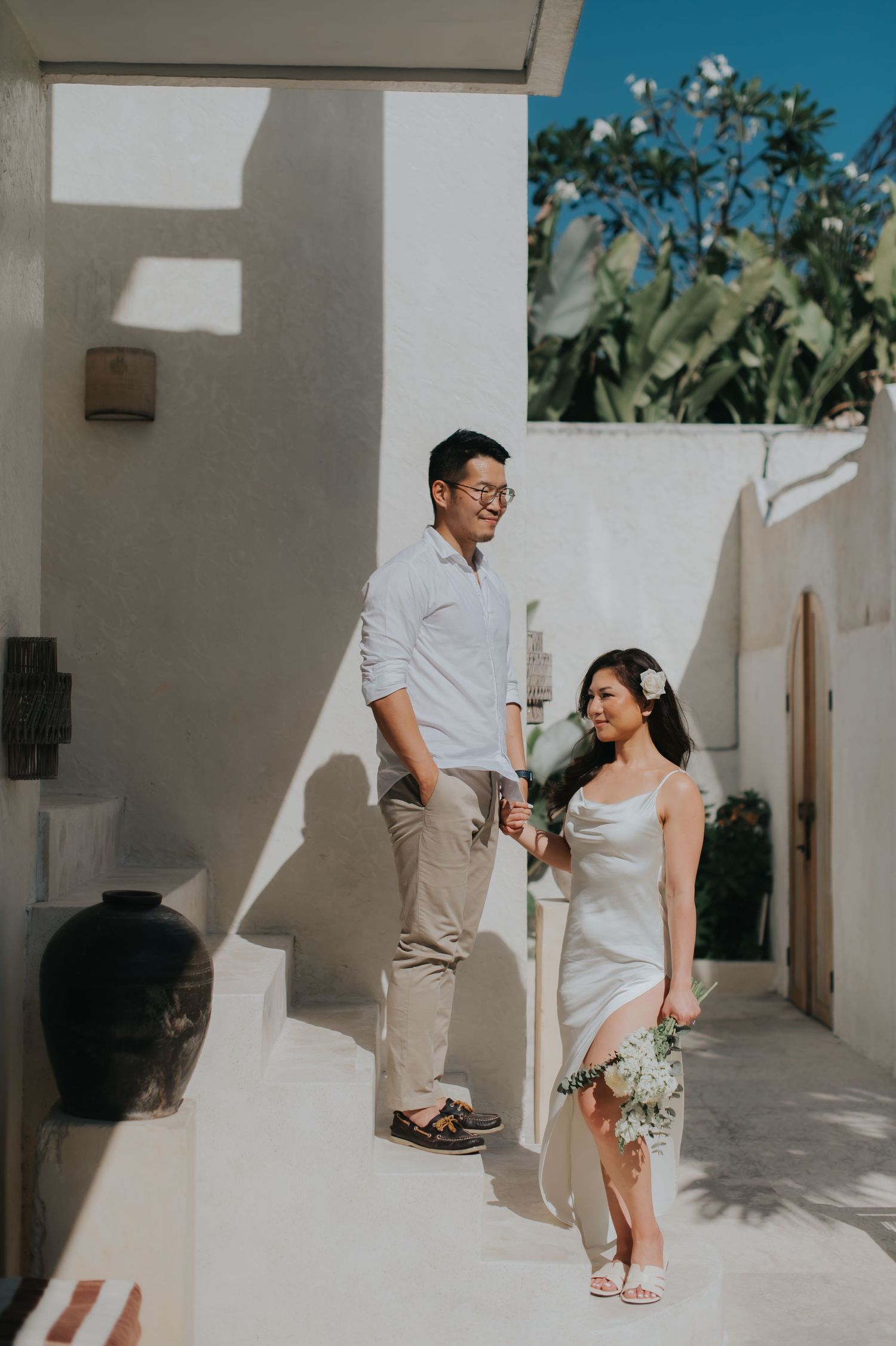 Couple in white outfits walking hand-in-hand on sunlit villa steps, woman holding bouquet with flower in hair.