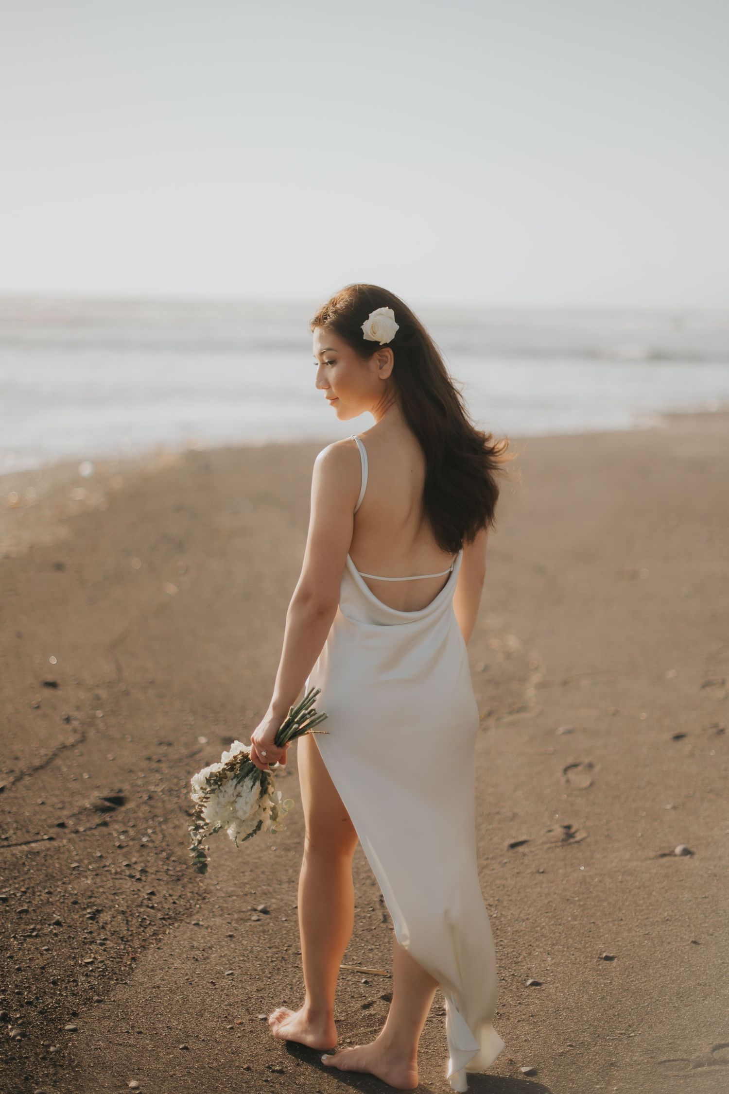 A bride in a white backless gown holds a bouquet while walking barefoot on a sandy beach at golden hour.