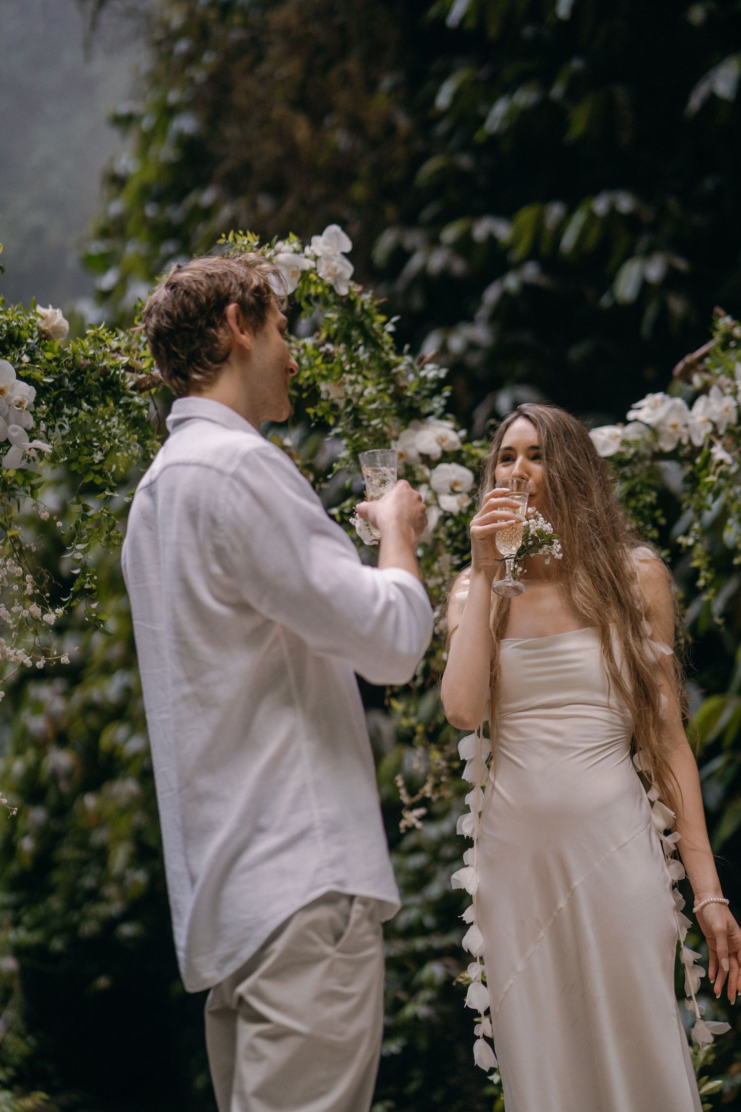 A couple in light-colored attire toasts with champagne glasses in a romantic garden setting surrounded by white flowers.