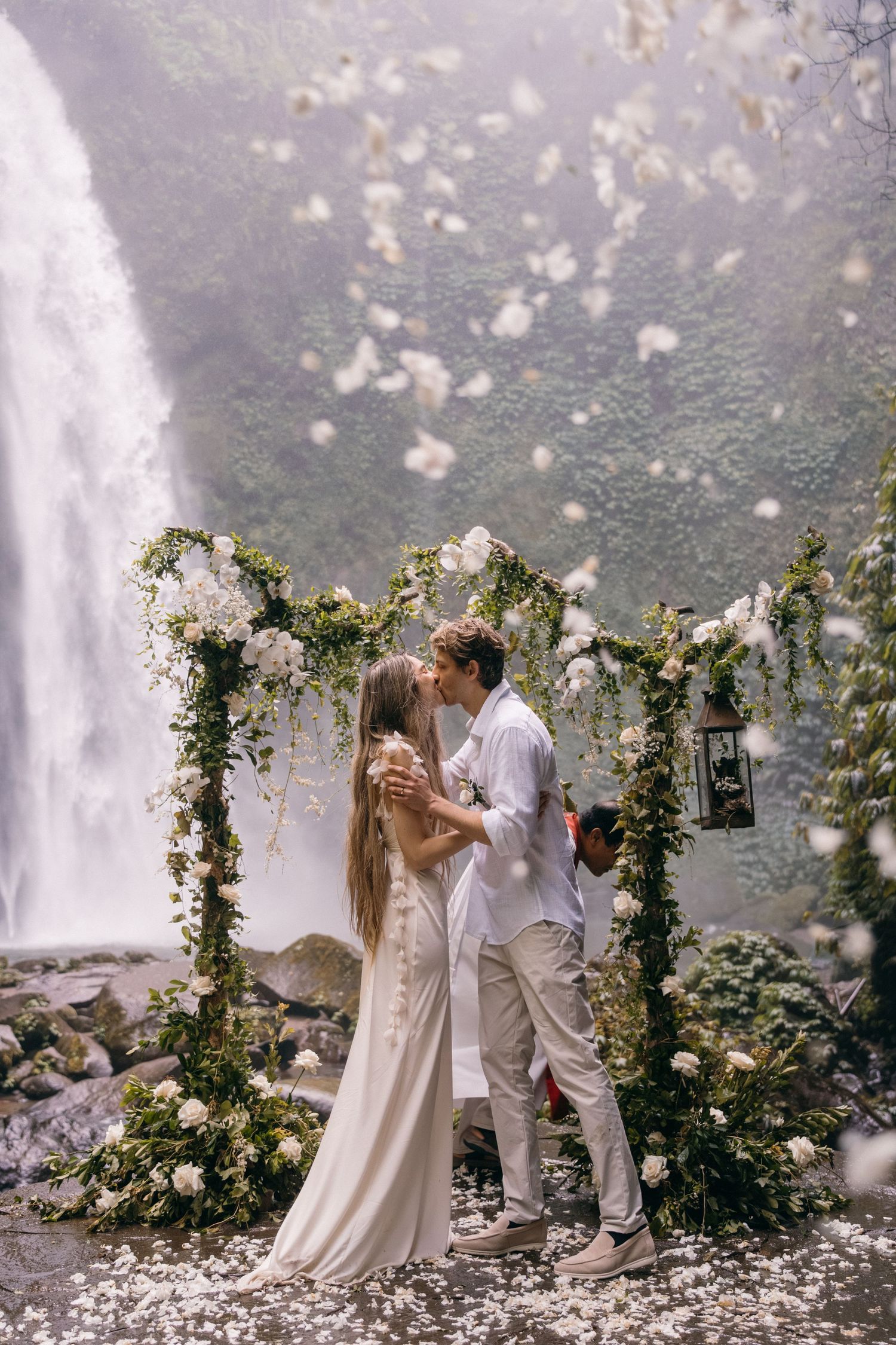 A couple shares a romantic moment under a floral arch by a waterfall as white petals fall around them in an ethereal scene.