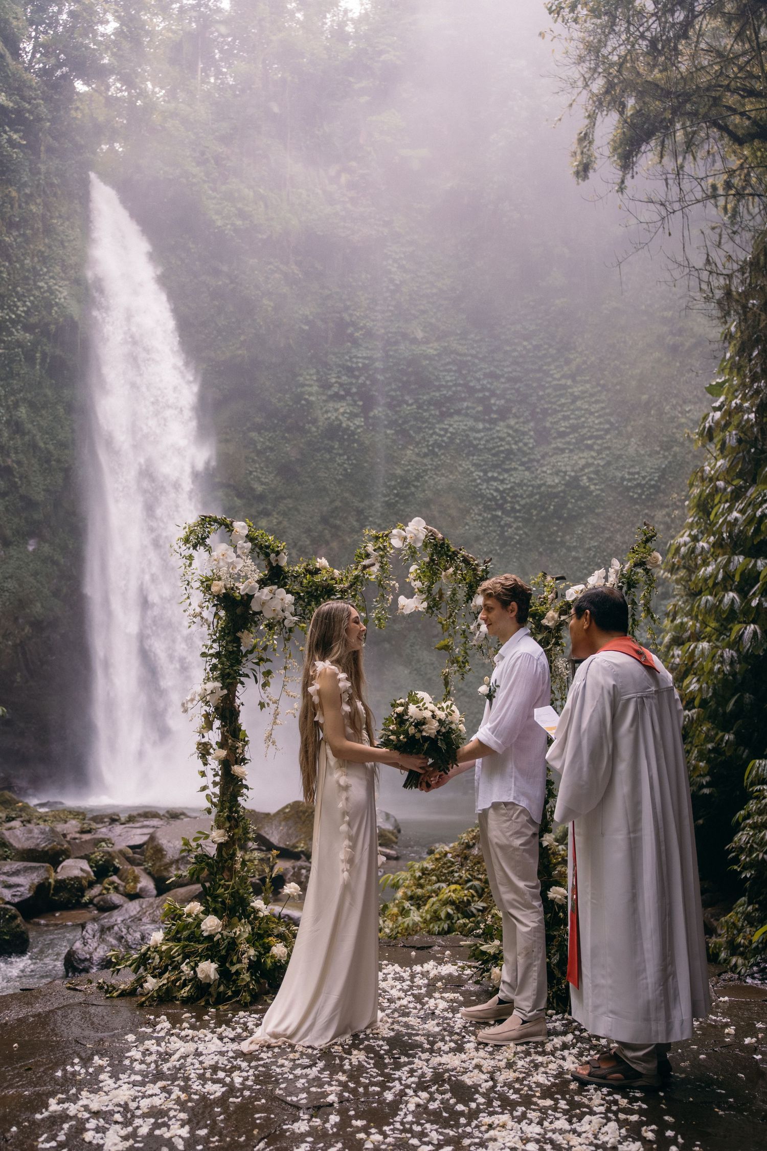 A wedding ceremony takes place in front of a misty waterfall with floral arches and natural decor in the forest.