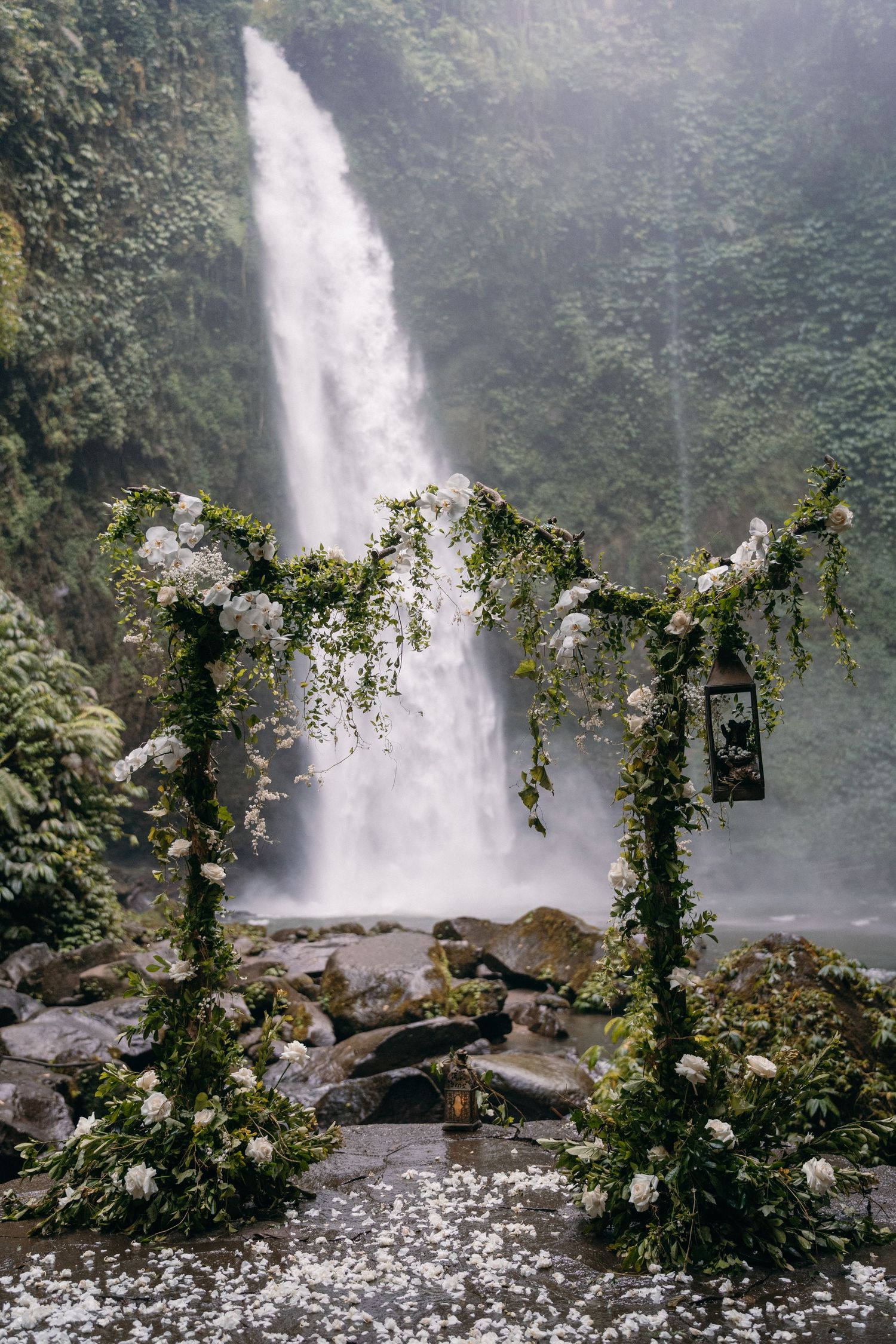 Majestic waterfall cascades behind floral archways with white roses and climbing vines over rocky terrain.