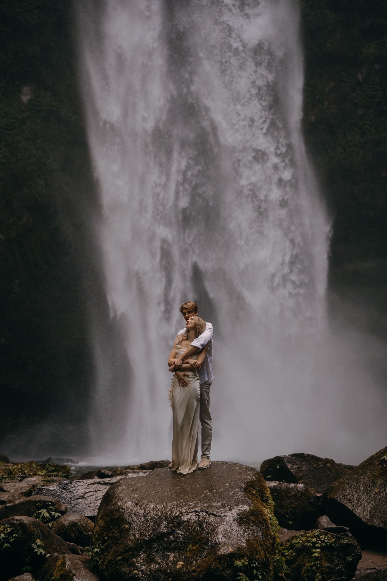 Couple embraces near majestic waterfall in moody atmospheric nature setting with dramatic mist and rocky landscape.