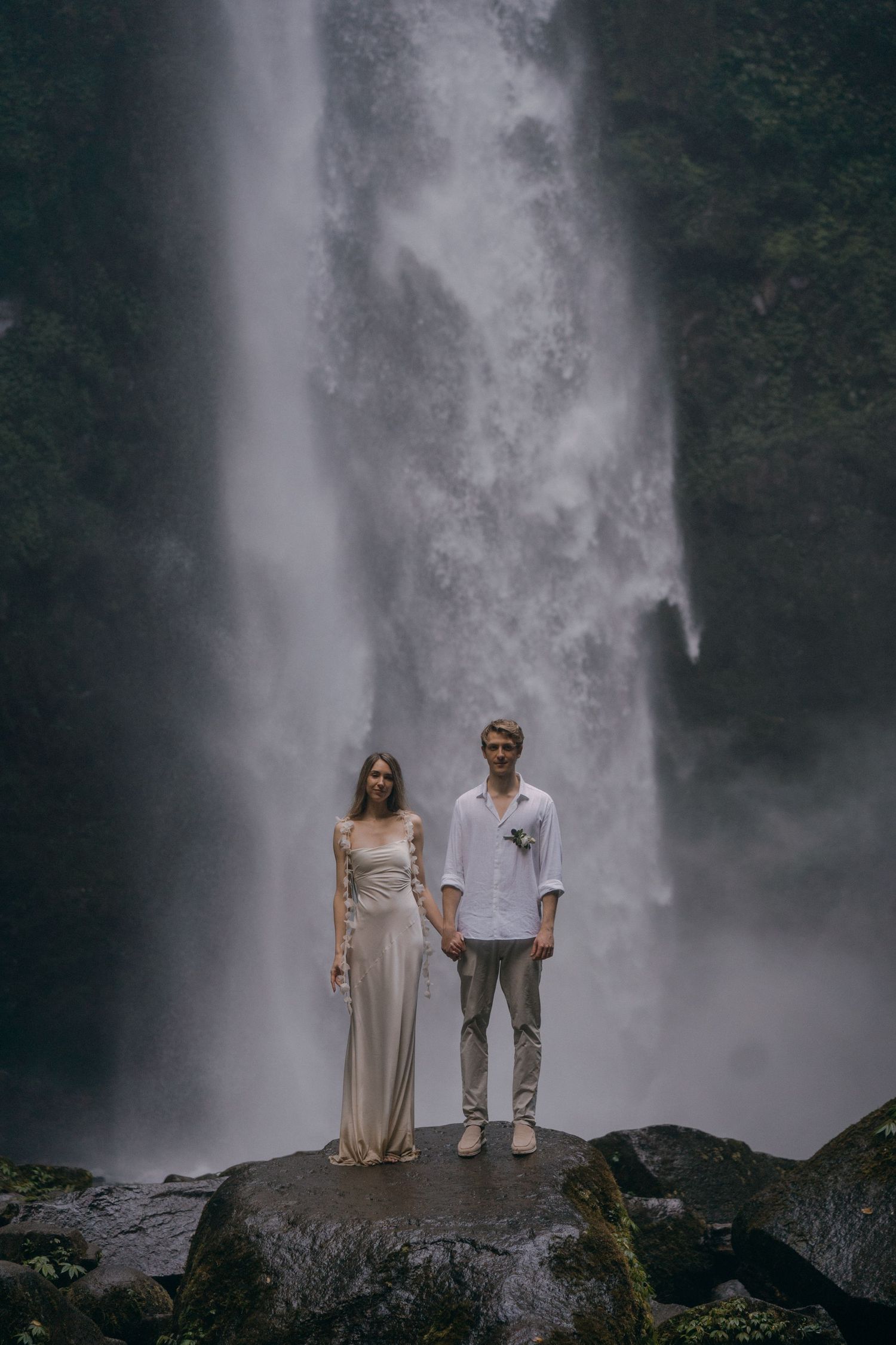 A couple stands on dark rocks in front of a majestic misty waterfall in an ethereal forest setting.