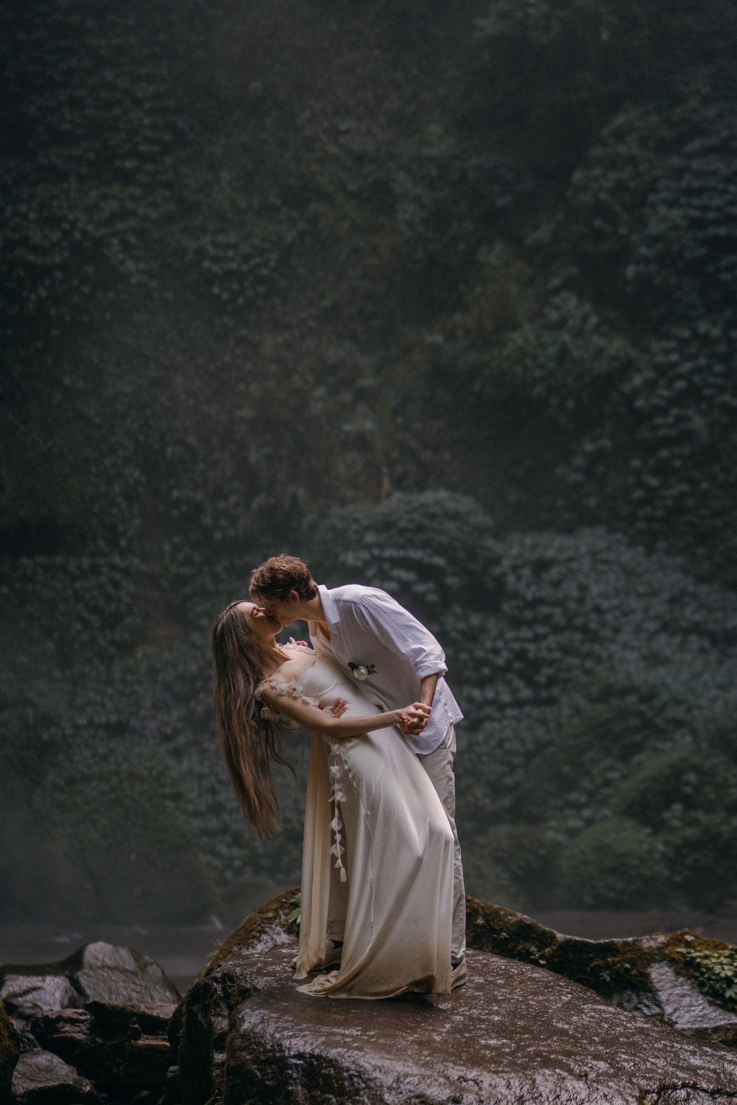 A romantic couple in white clothing embraces while sitting on a rocky cliff edge at dusk.