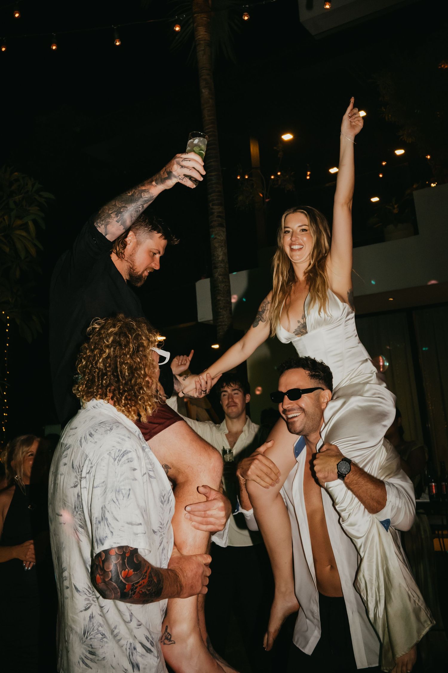Excited bride raised on guests shoulders while groom celebrates at lively tropical wedding reception.