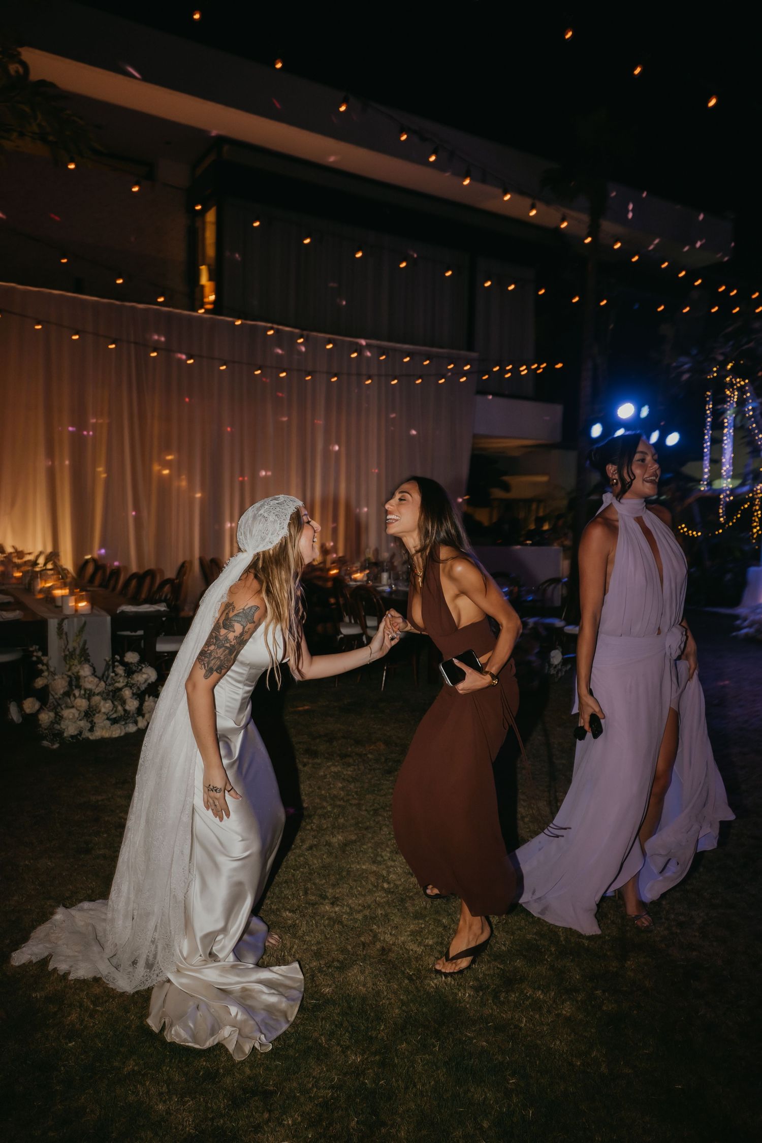 Bride in white gown dancing joyfully with guests at an outdoor evening wedding reception with string lights.