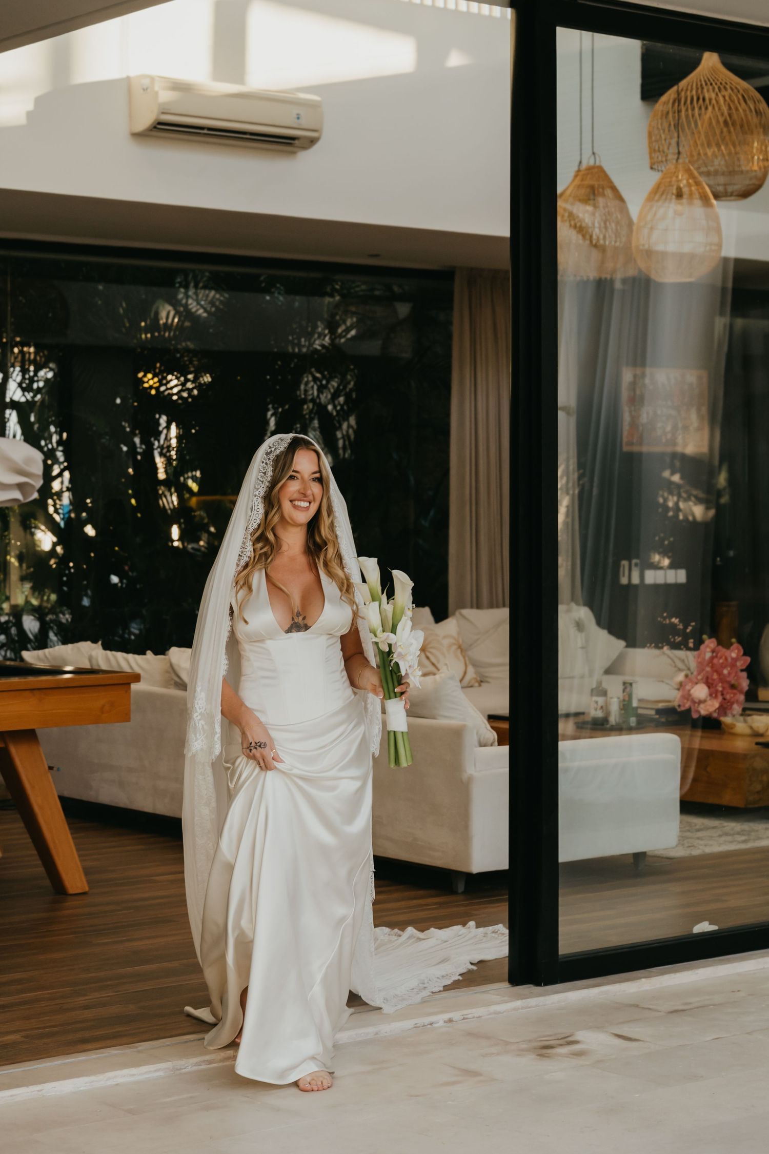 Smiling bride in fitted white gown and lace veil holding calla lily bouquet in modern villa.