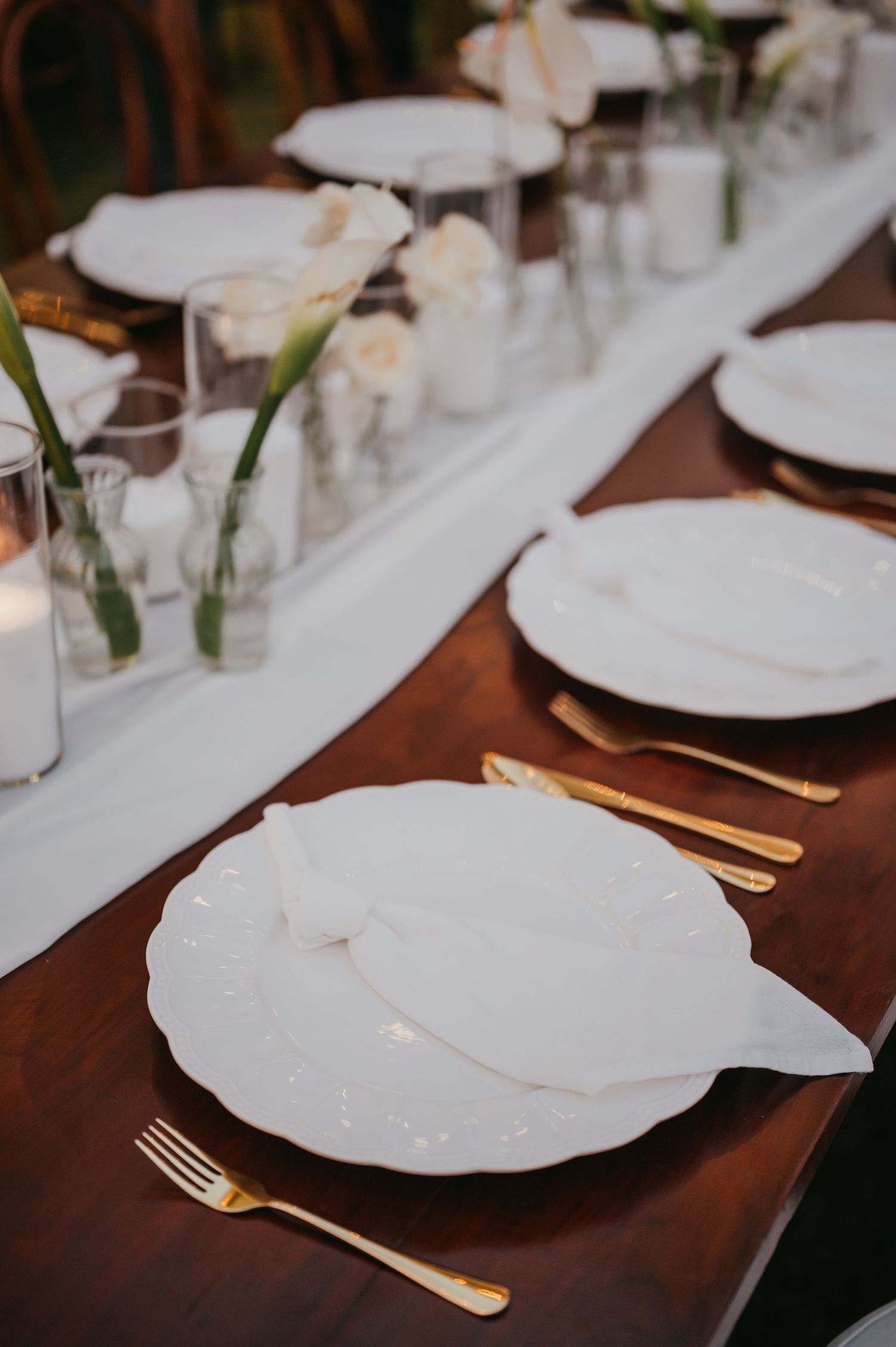 Elegant wedding reception table setting with white plates, gold cutlery, and white floral centerpieces.