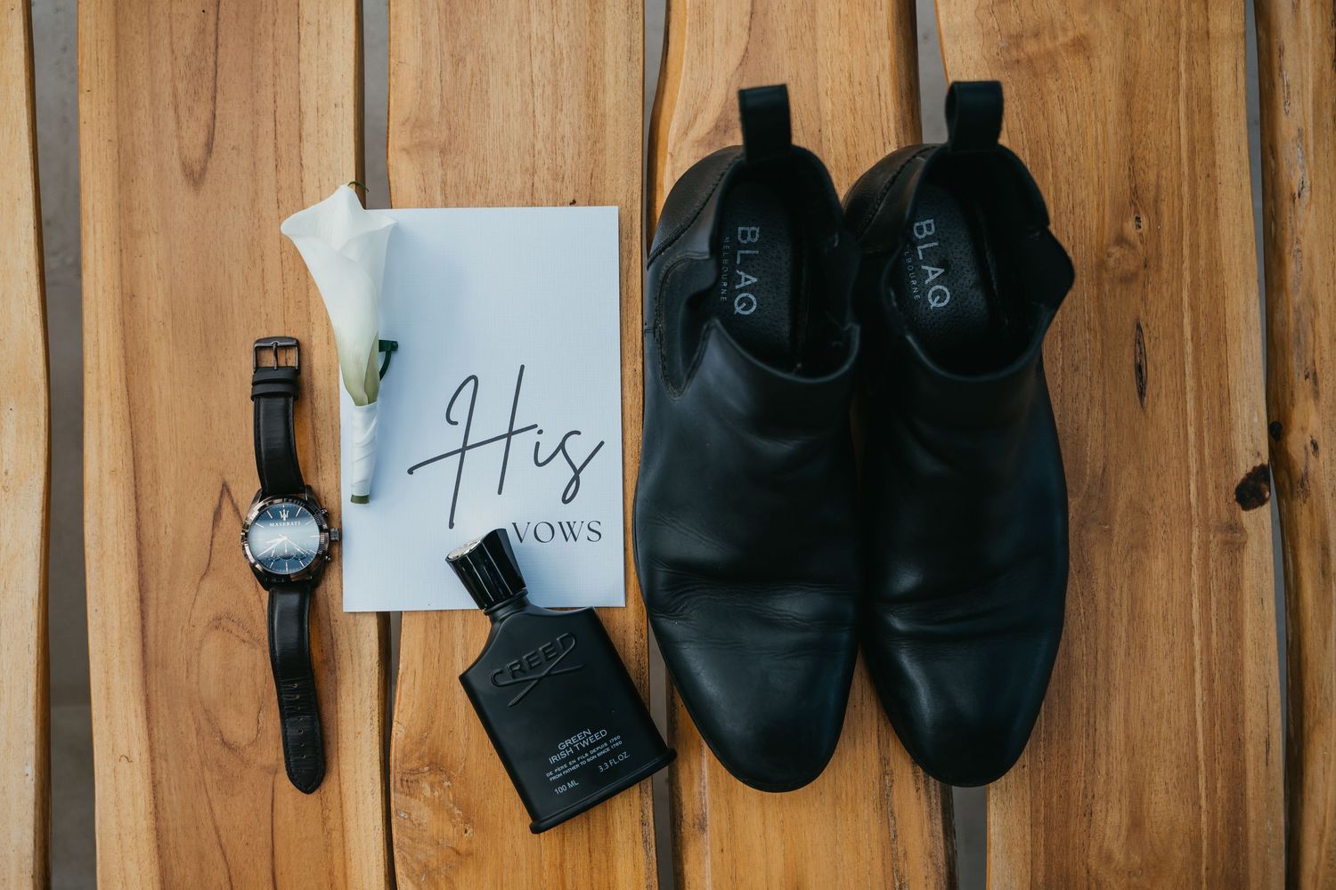 Black leather Chelsea boots, watch, cologne, and His Vows booklet arranged on wooden surface for groom wedding flat lay.
