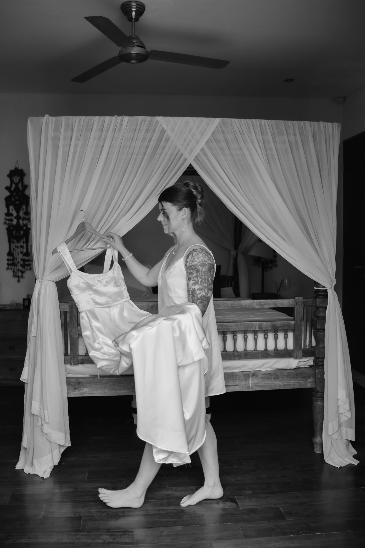Black and white photo of tattooed bride holding wedding dress near canopy bed in villa.