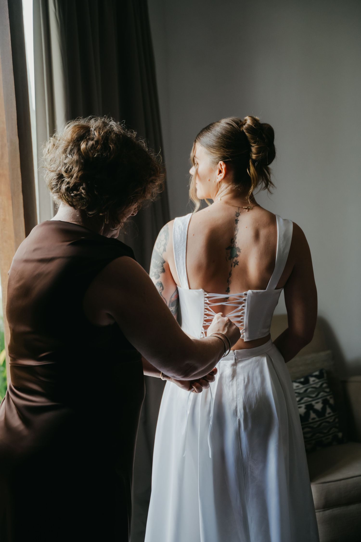 Mother helps lace up the back of brides elegant white wedding gown in soft natural window light.