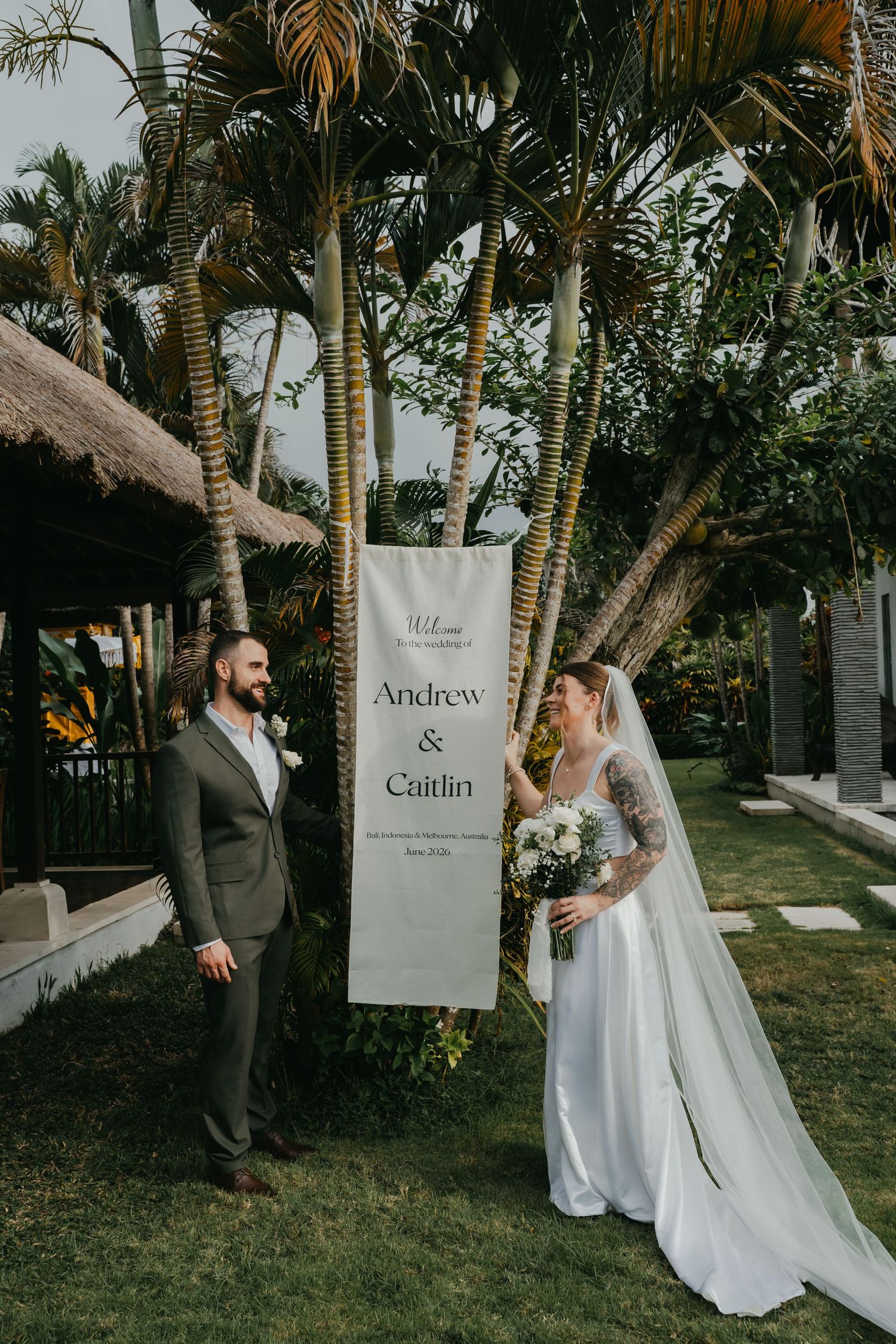 Bride and groom pose beside Welcome Andrew and Caitlin wedding banner among tropical palm trees.