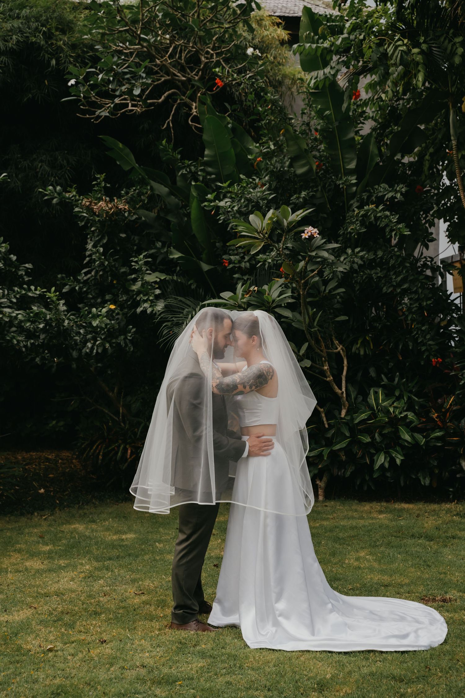 Bride and groom share a kiss hidden beneath wedding veil in front of tropical greenery.