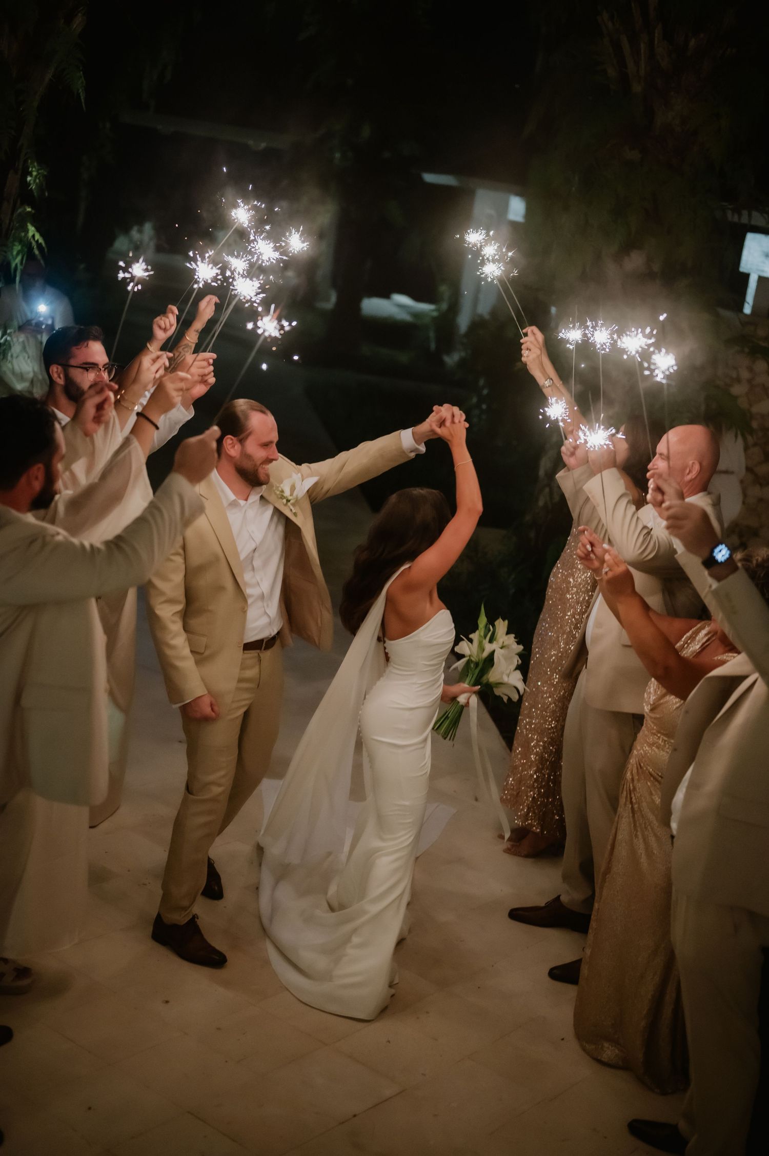 Newlyweds walk through sparkler tunnel with wedding party during celebratory nighttime send-off.