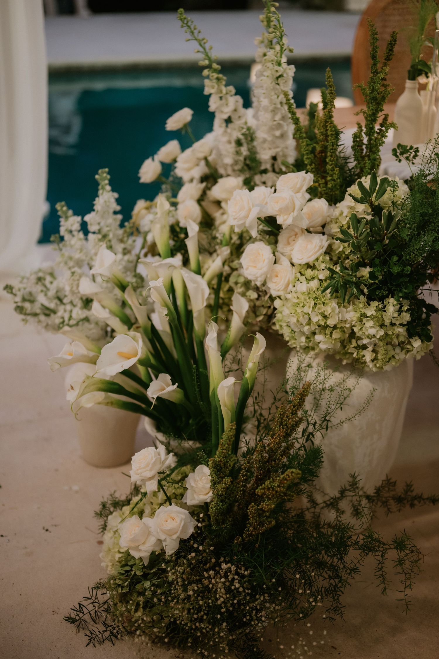 Elegant white wedding floral arrangements with calla lilies and roses beside a blue pool.