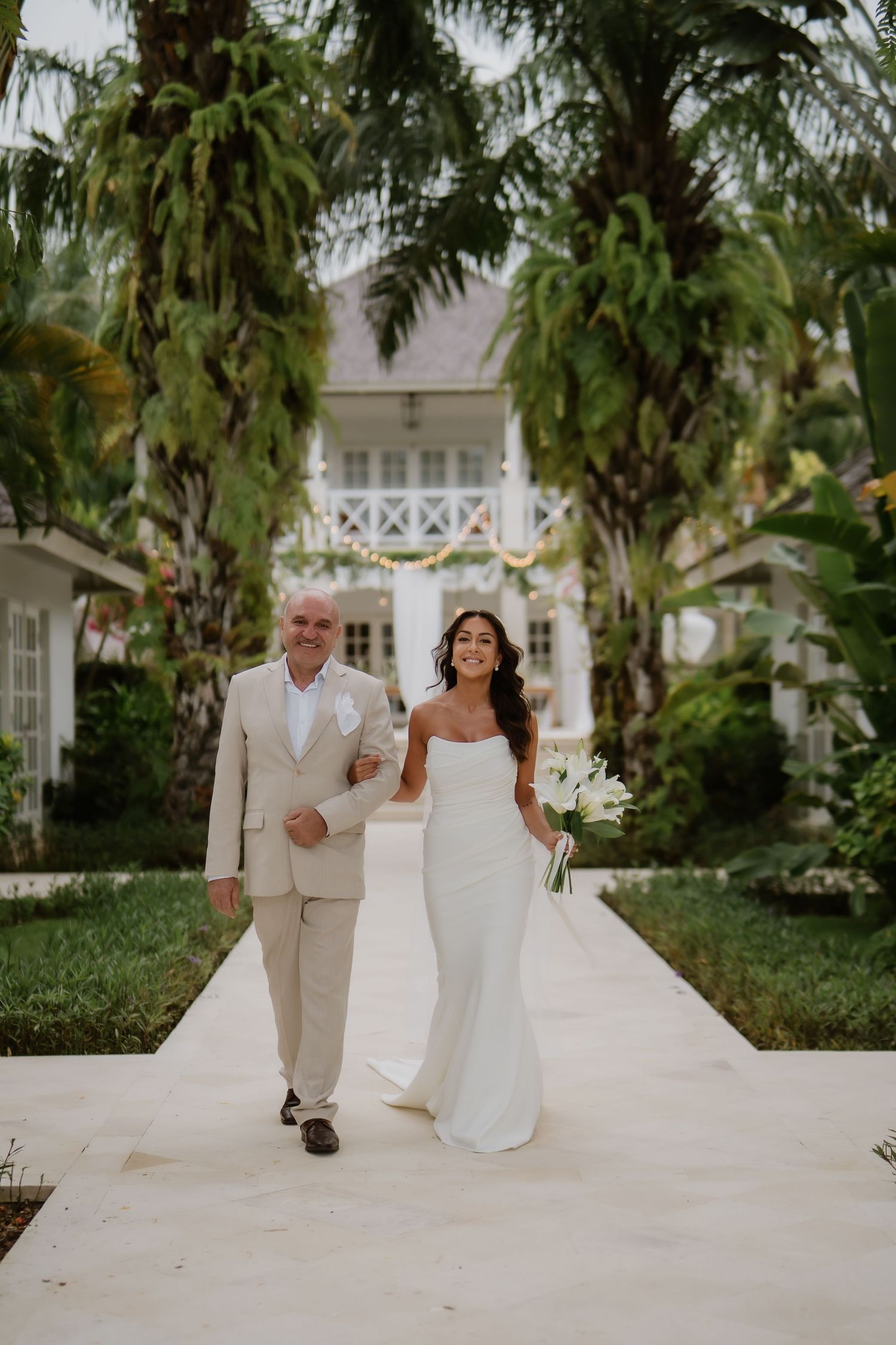 Bride carrying white calla lily bouquet walking arm-in-arm with father down palm-lined wedding aisle.