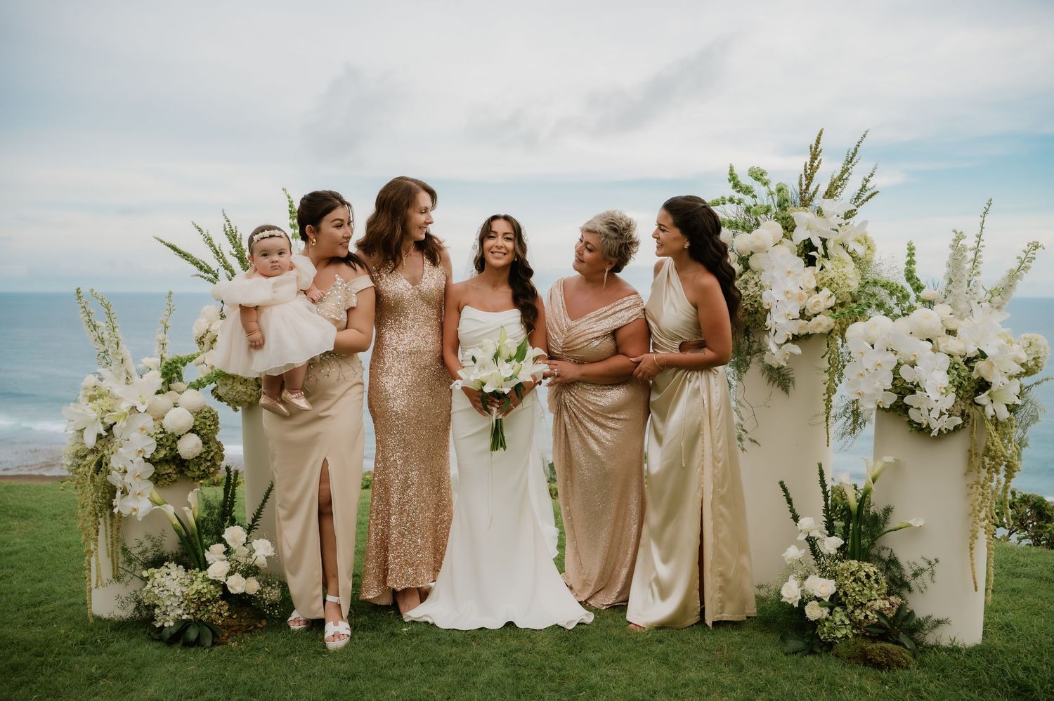 Bride with bridesmaids in gold dresses pose at oceanfront floral wedding altar.