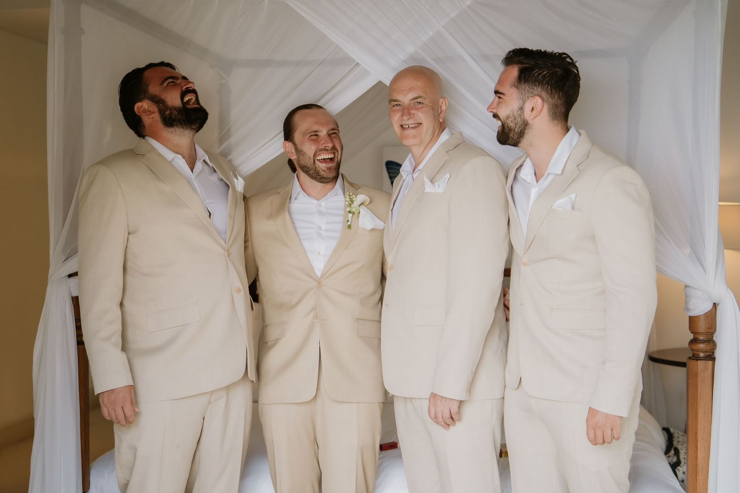 Four groomsmen in matching tan suits laughing together before a wedding ceremony.