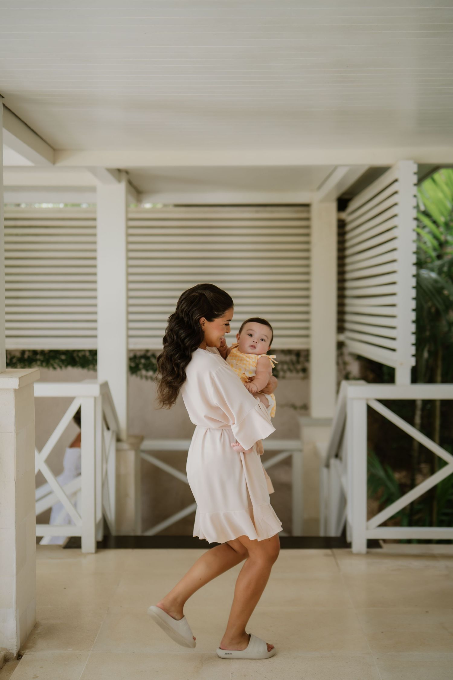 A woman in a white robe holds a baby on a tropical resort veranda.