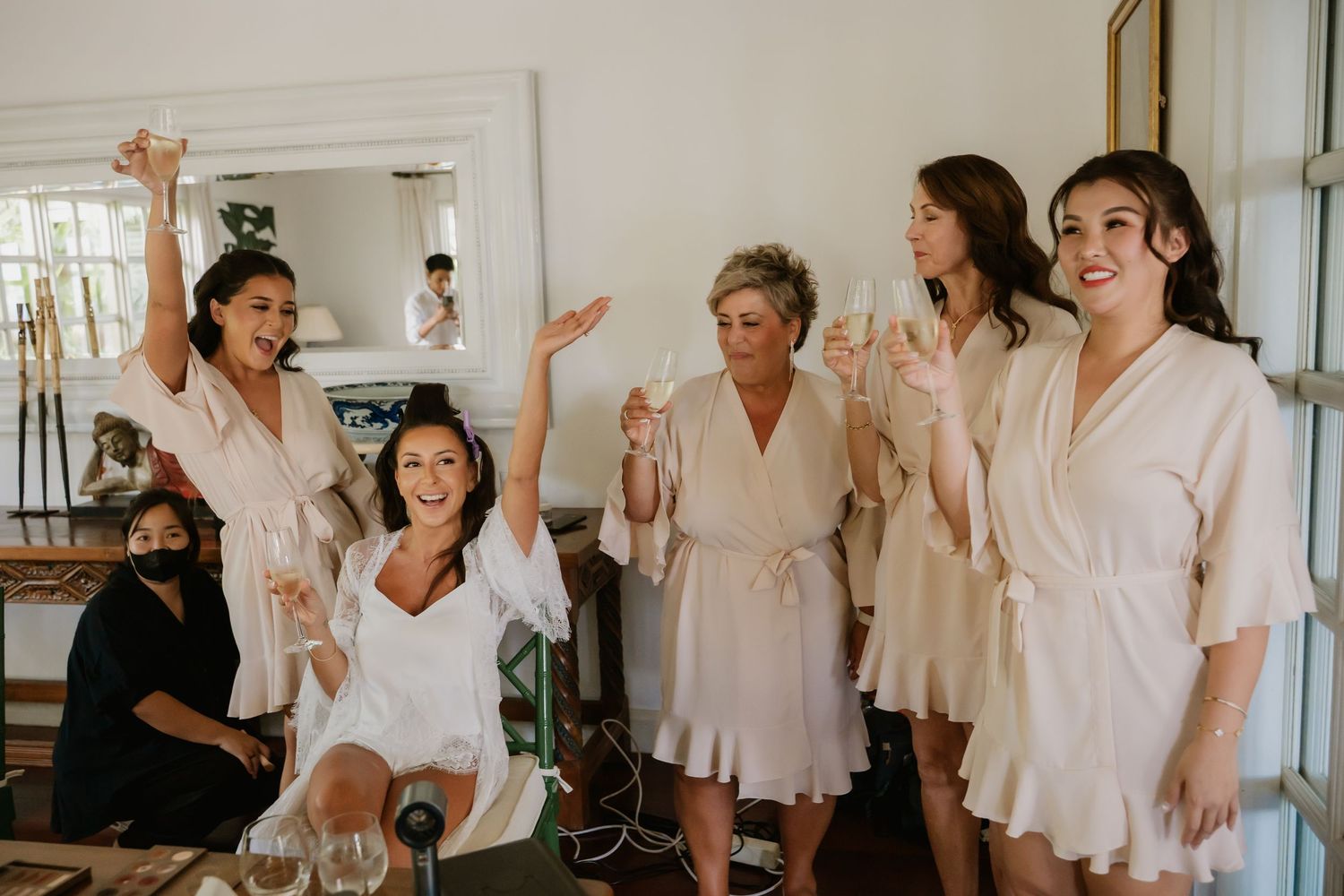 Bridesmaids in blush robes celebrate with champagne during wedding morning preparations.