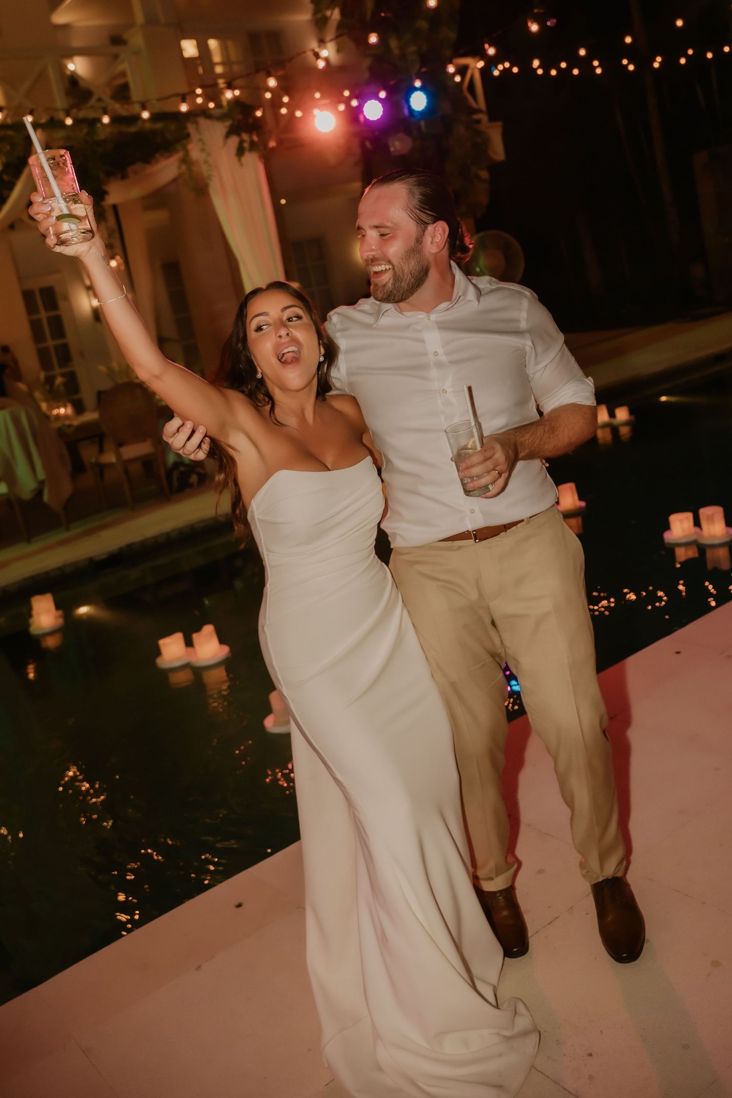 Joyful bride in white strapless gown and groom celebrate with drinks beside a pool at night.