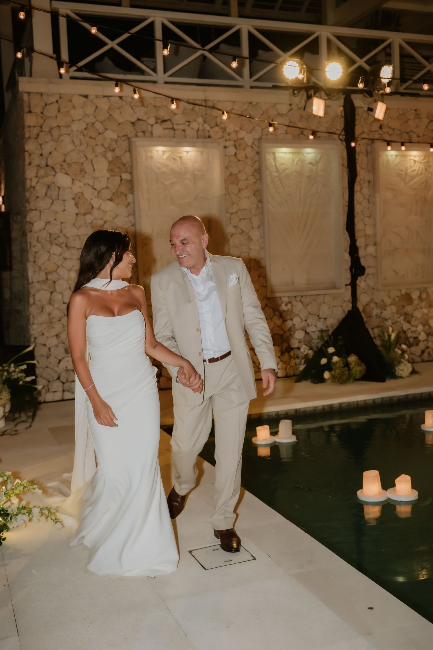 Bride and older man in beige suit walk hand in hand beside candle-lit pool at night.